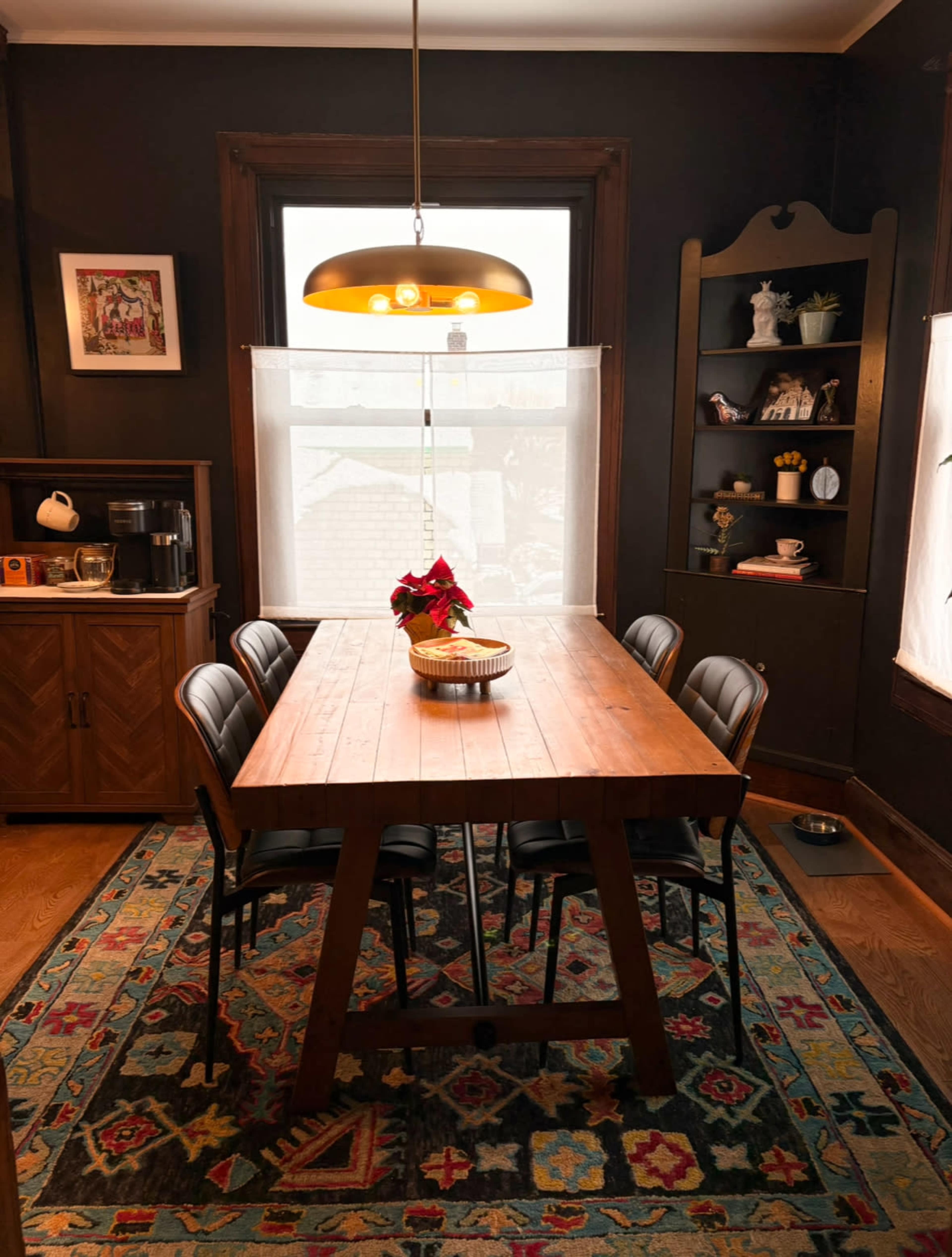 The image shows a dining room with a wooden table surrounded by black chairs, a decorative rug, and a window with light filtering through sheer curtains.