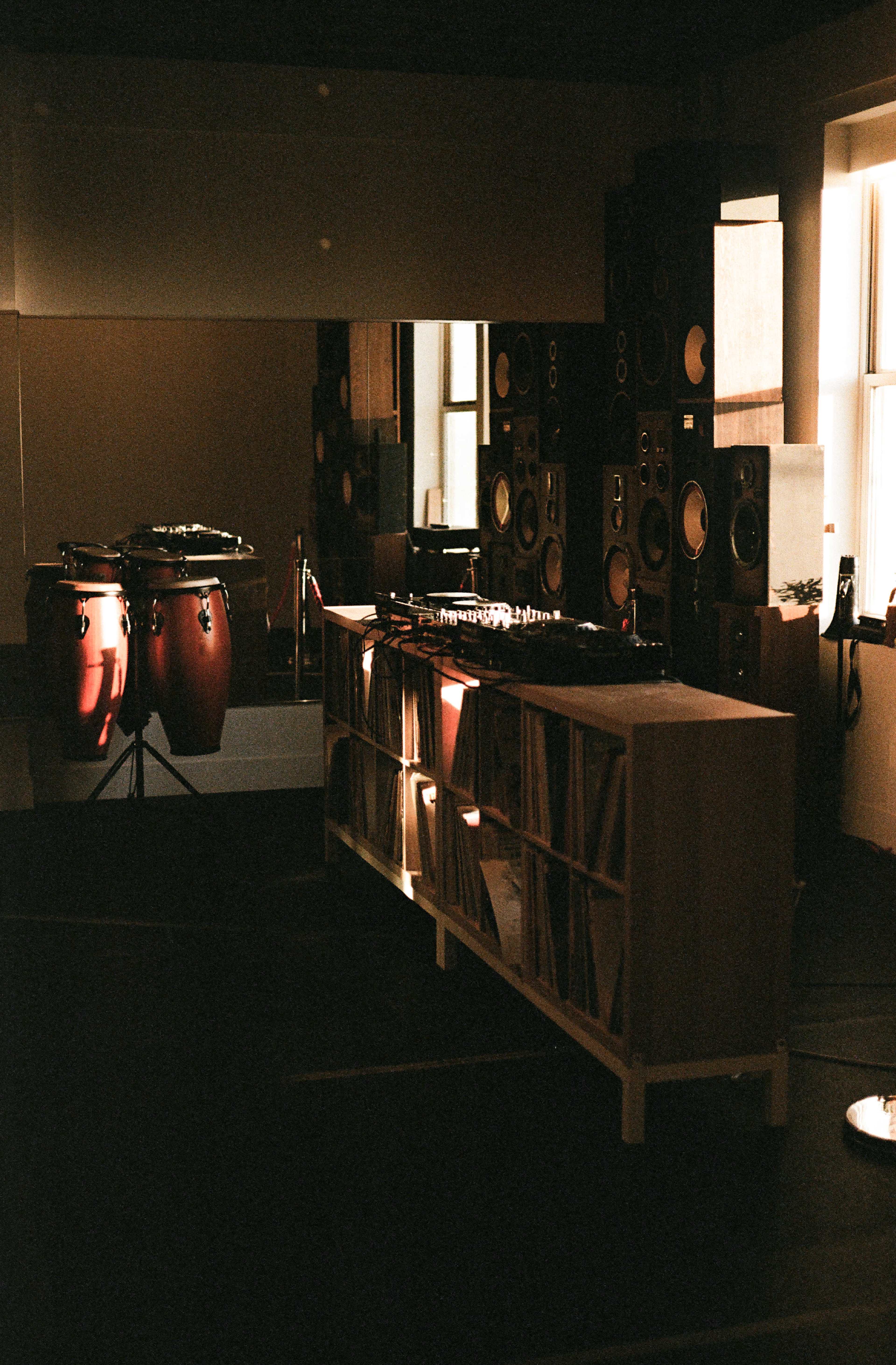 The image shows a music studio featuring a DJ setup with turntables and vinyl records, surrounded by large speakers and conga drums, illuminated by natural light.