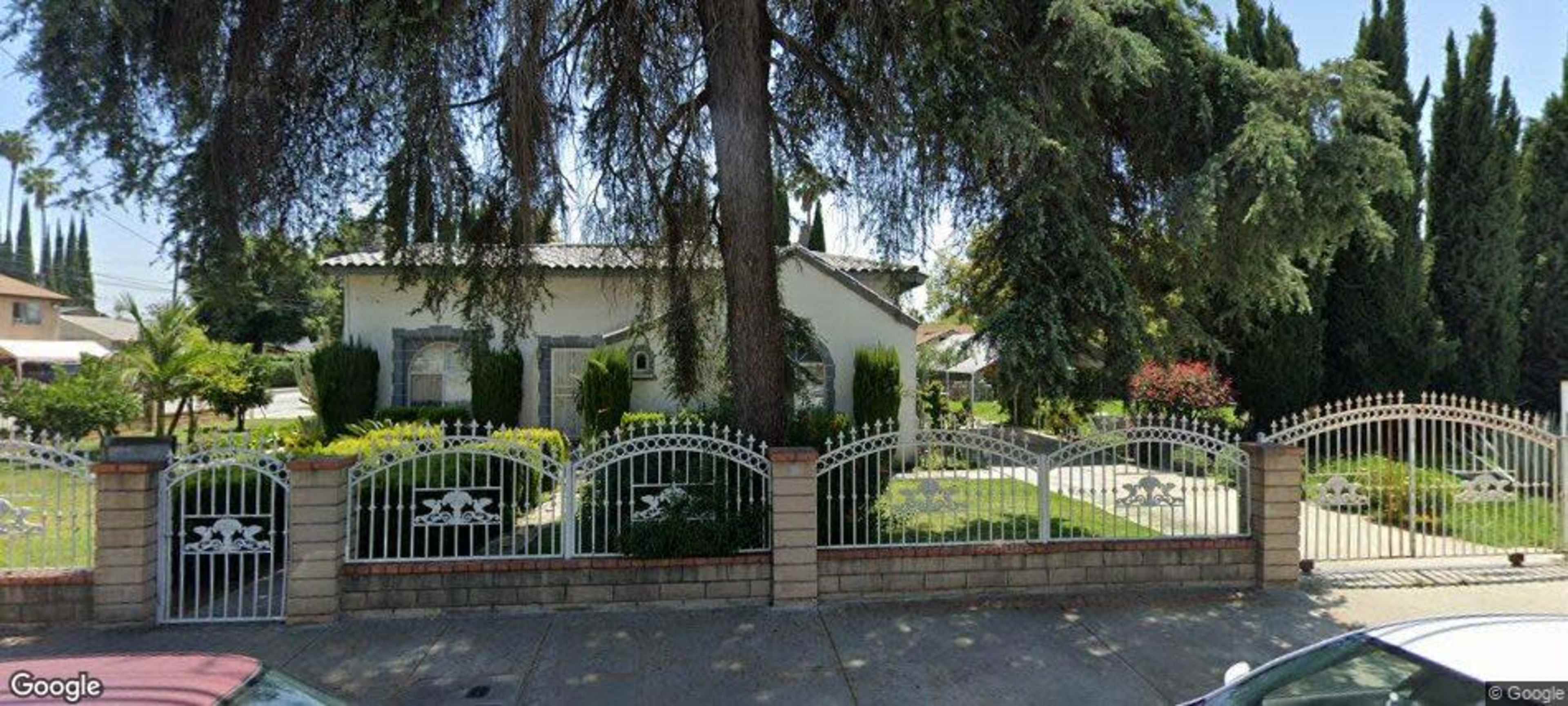 The image shows a suburban house surrounded by a decorative gate and well-maintained landscaping with various trees and plants.