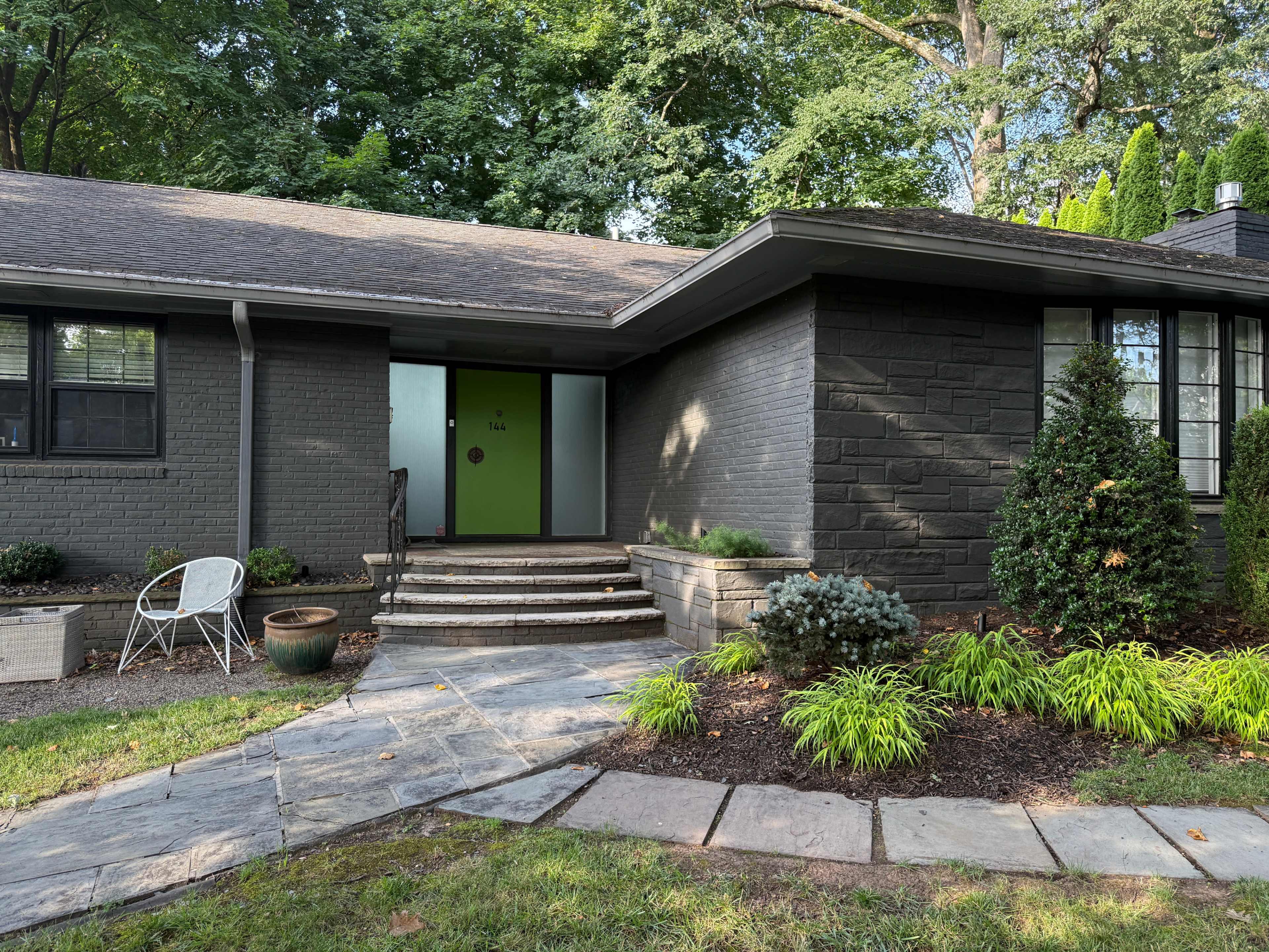 A single-story house with a gray exterior features a set of stone steps leading to a glass front door, surrounded by landscaped greenery.