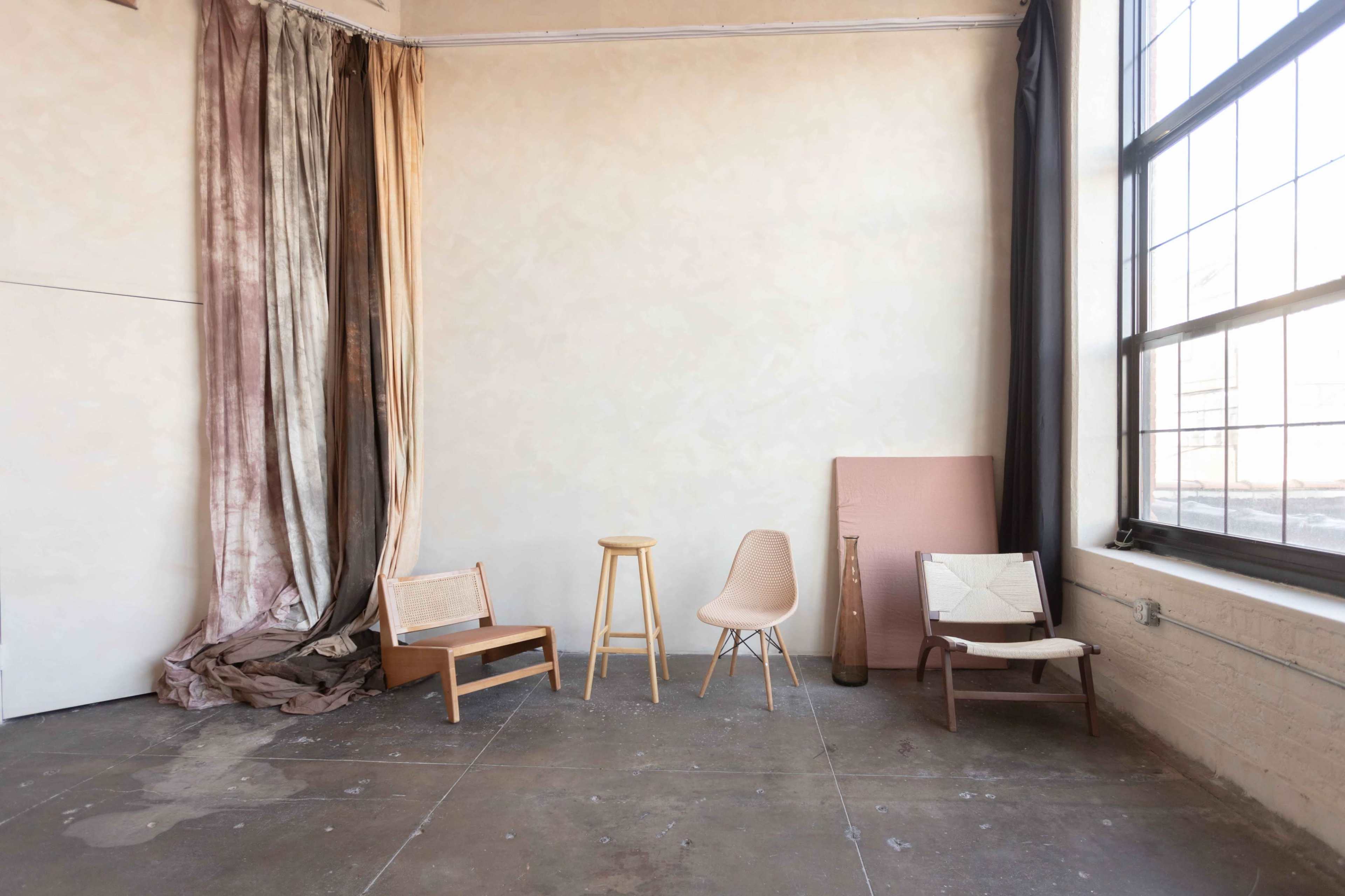 A spacious interior with a bare concrete floor, two chairs, a stool, and draped fabric near a large window.