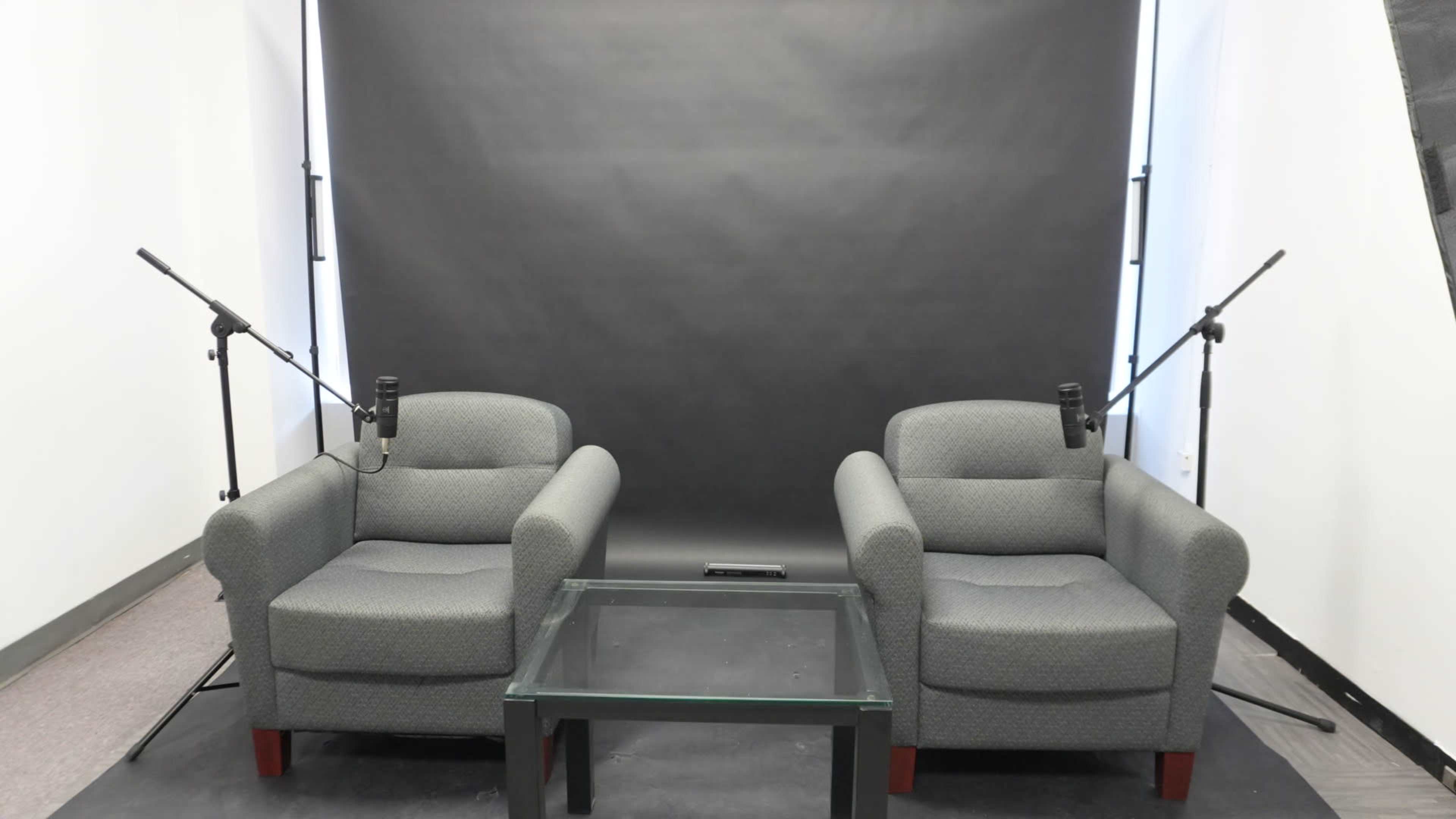 The scene features two gray armchairs facing each other over a glass table, with microphones positioned on either side in front of a black backdrop.