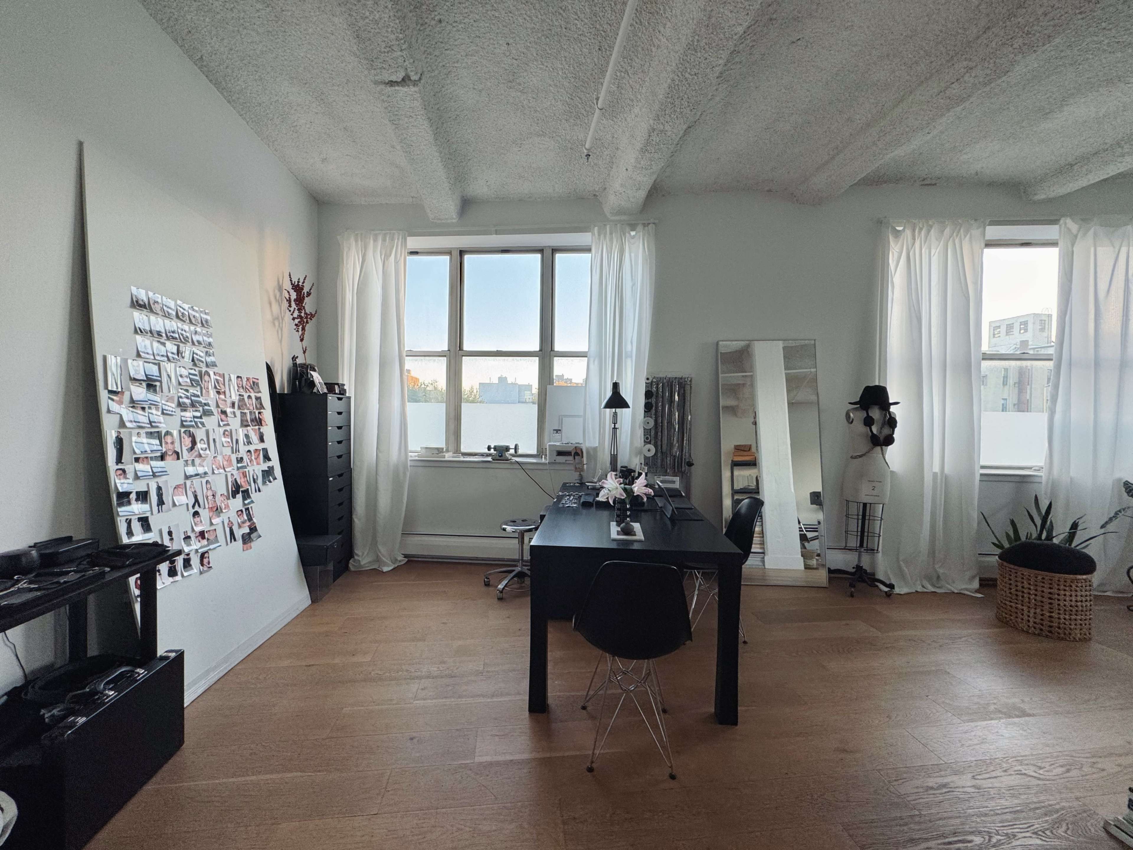 The image shows a spacious, well-lit room featuring a black desk with a chair, a large mirror, a wall adorned with photographs, and several windows with sheer white curtains.