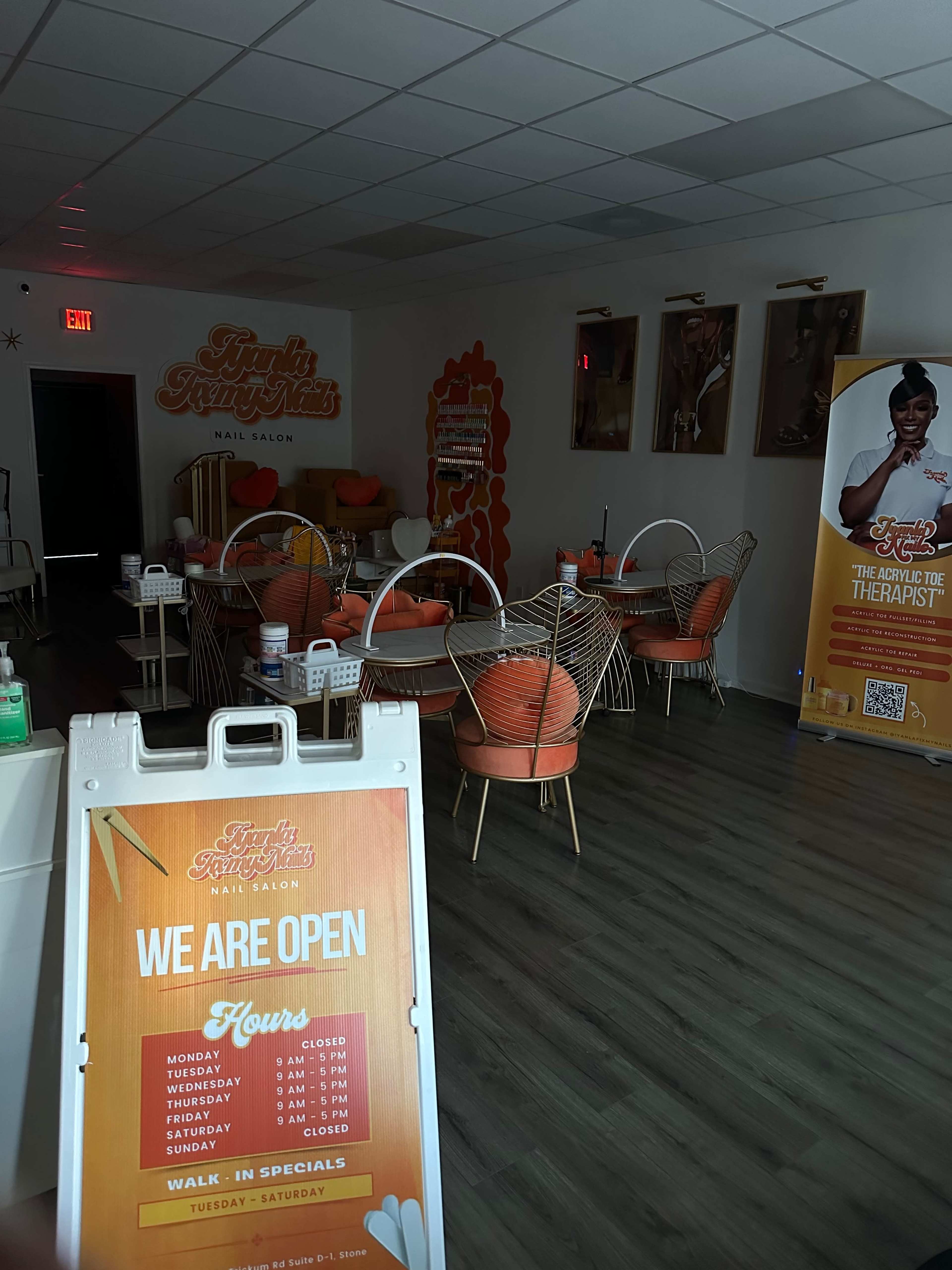 The interior of a nail salon with orange decor, featuring seating and a sign indicating business hours.