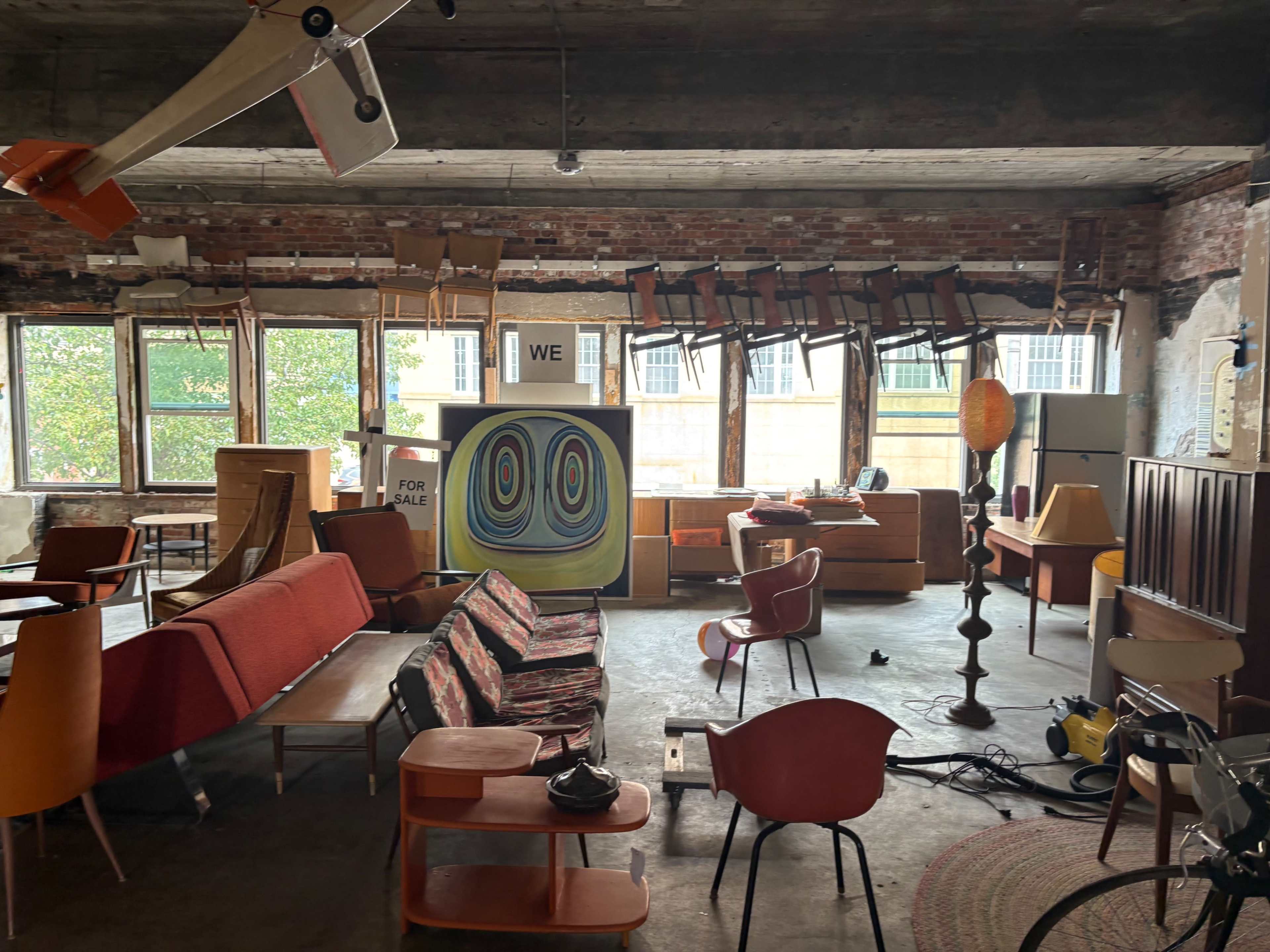 The interior of a vintage furniture store features various retro seating arrangements, stacked chairs on the ceiling, and colorful artwork on the walls.