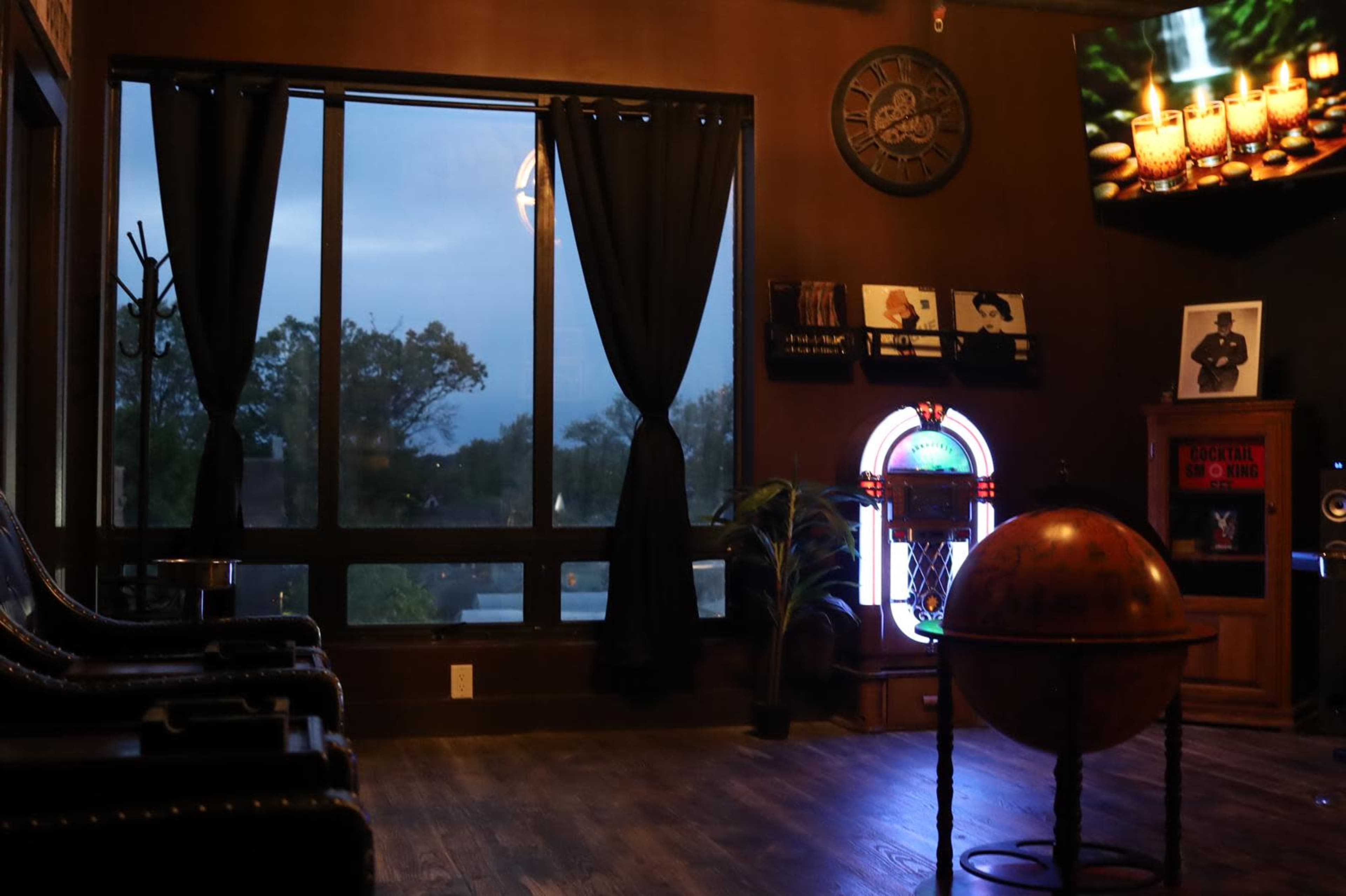 The image shows an interior space with dark wood flooring, large windows covered by black curtains, and a decorative globe on a table near a stained glass piece and various artworks on the walls.