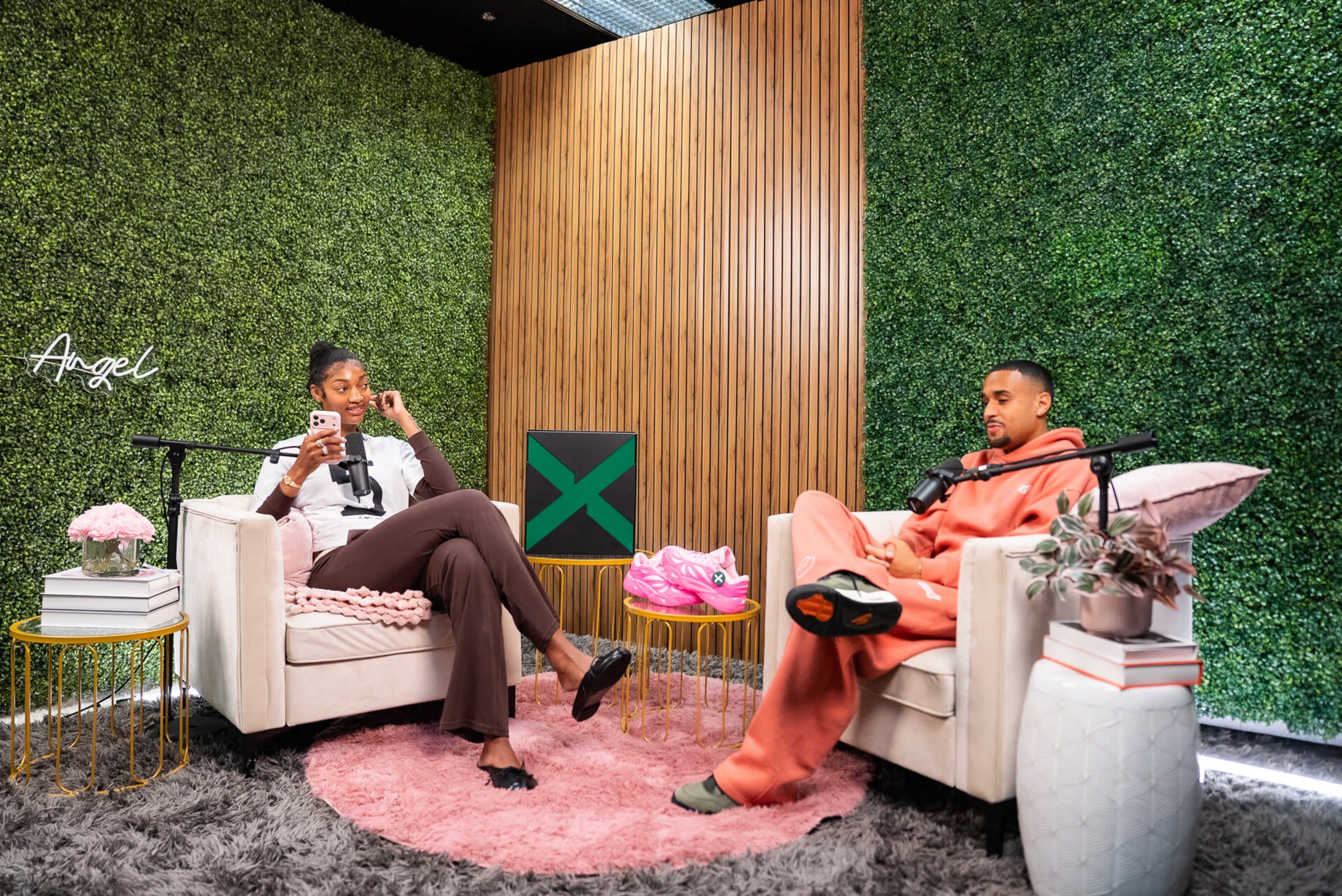 Two people are seated on white couches in a green-walled studio, with one person checking their phone while the other speaks into a microphone.