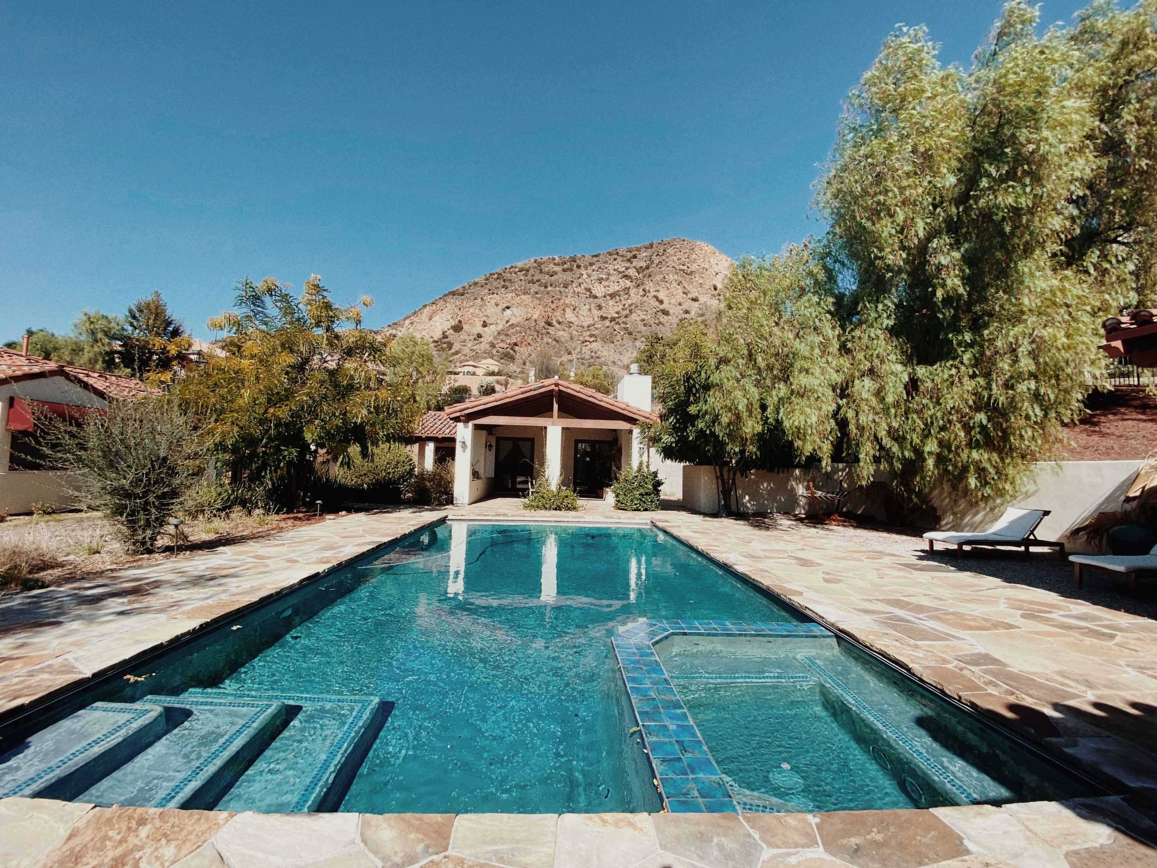 A rectangular swimming pool with a hot tub is surrounded by stone tiles and landscaping, set against a backdrop of a mountain on a clear day.
