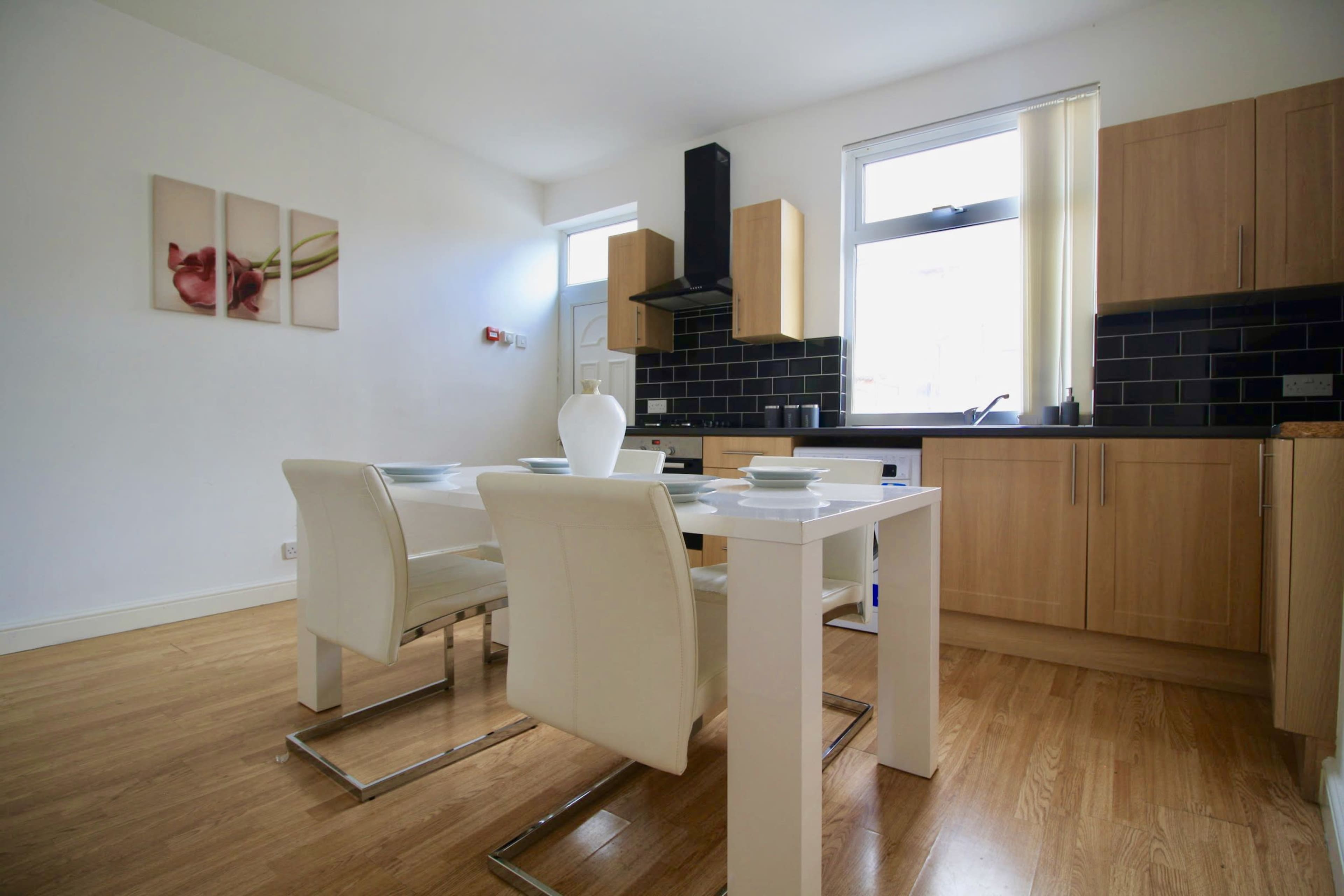 A modern kitchen features a wooden dining table with white chairs, sleek cabinetry, and black tiled walls.