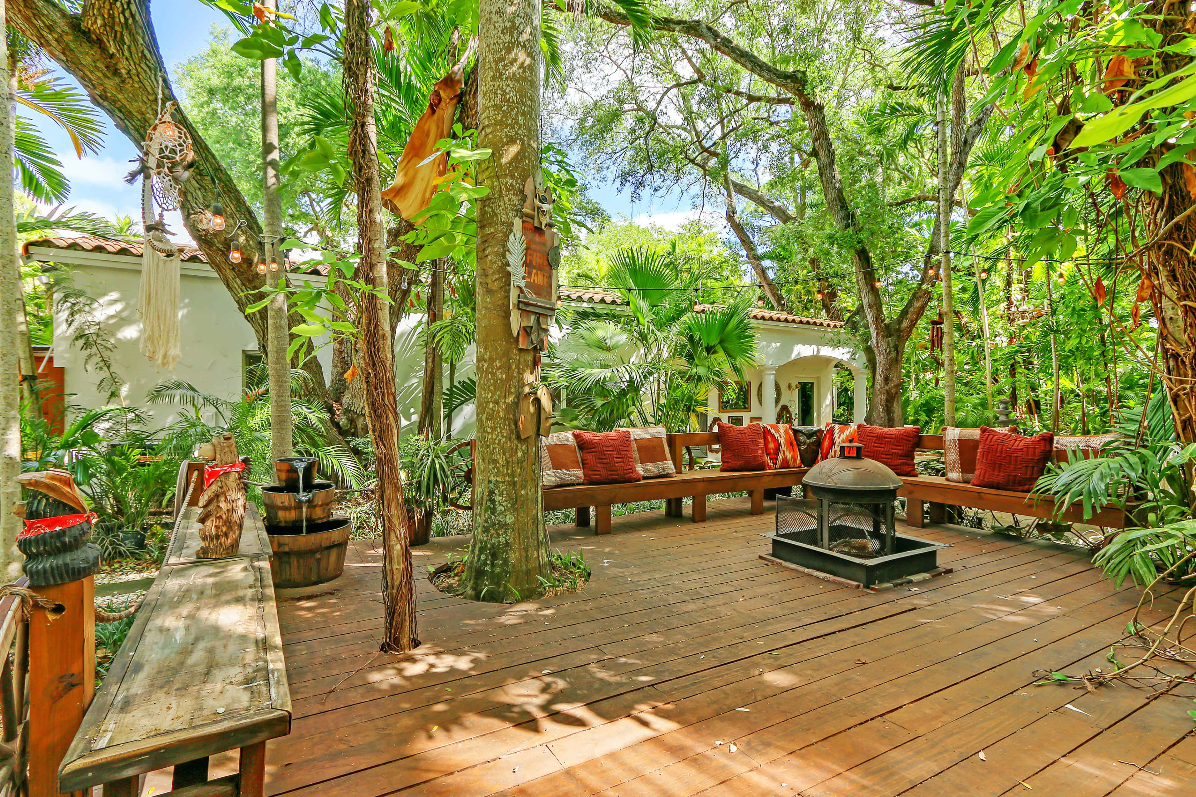 A wooden deck surrounded by lush greenery, featuring a seating area with cushions and a central fire pit.