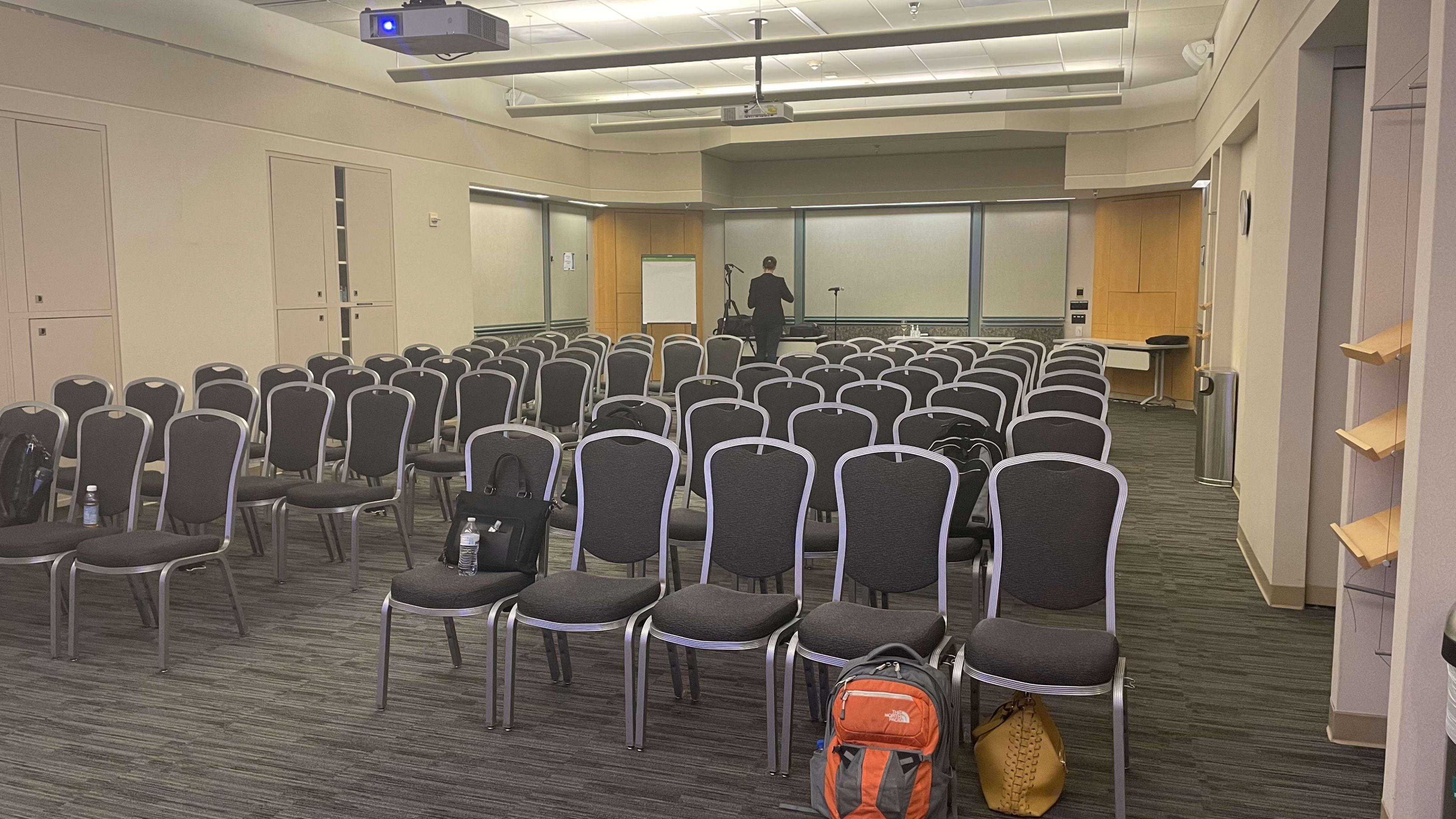 A conference room with rows of empty chairs and a person setting up a presentation at the front.