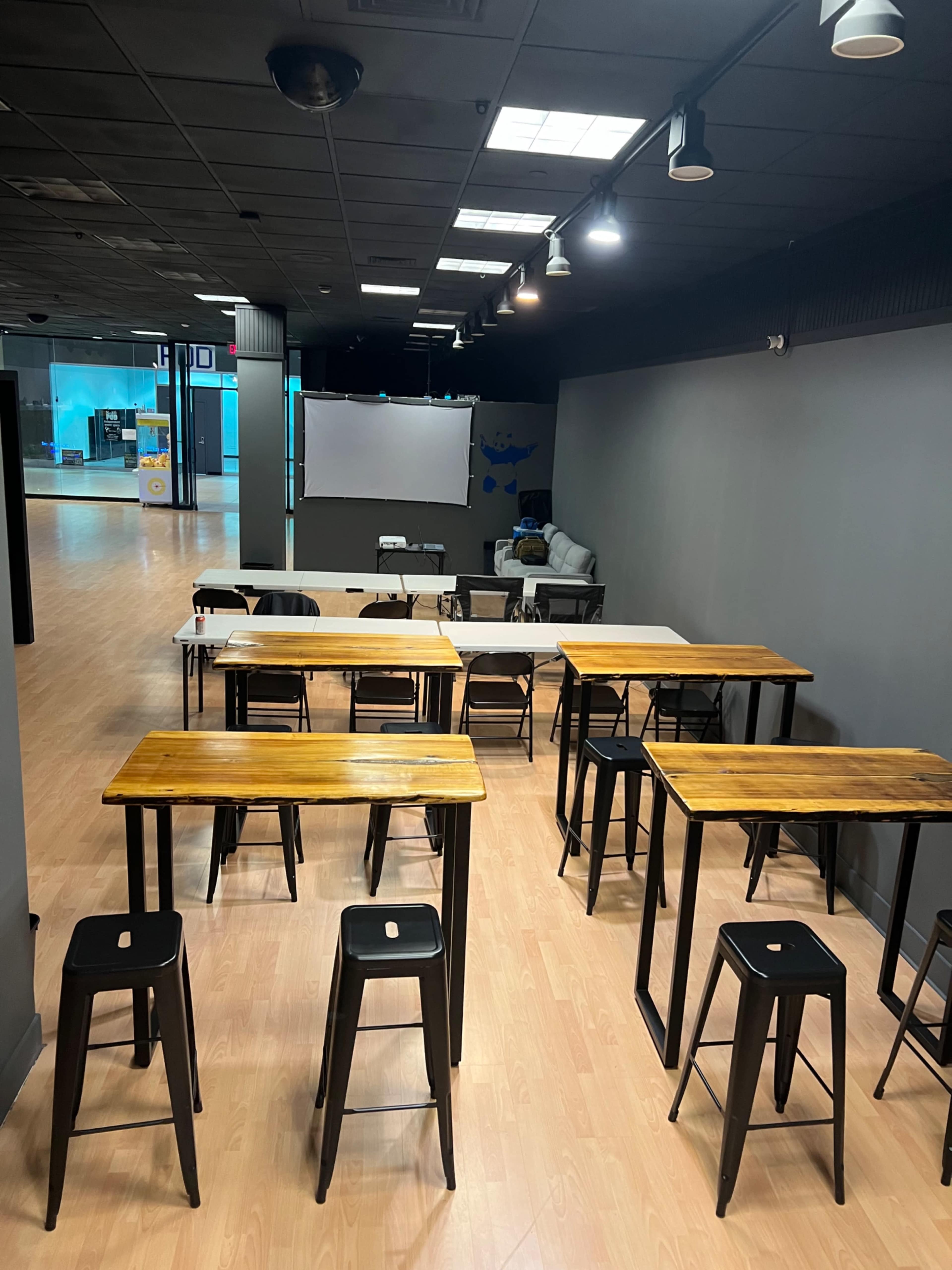 The image shows a spacious room with several wooden tables and black stools arranged in a layout, alongside a blank projection screen and a couch in the background.