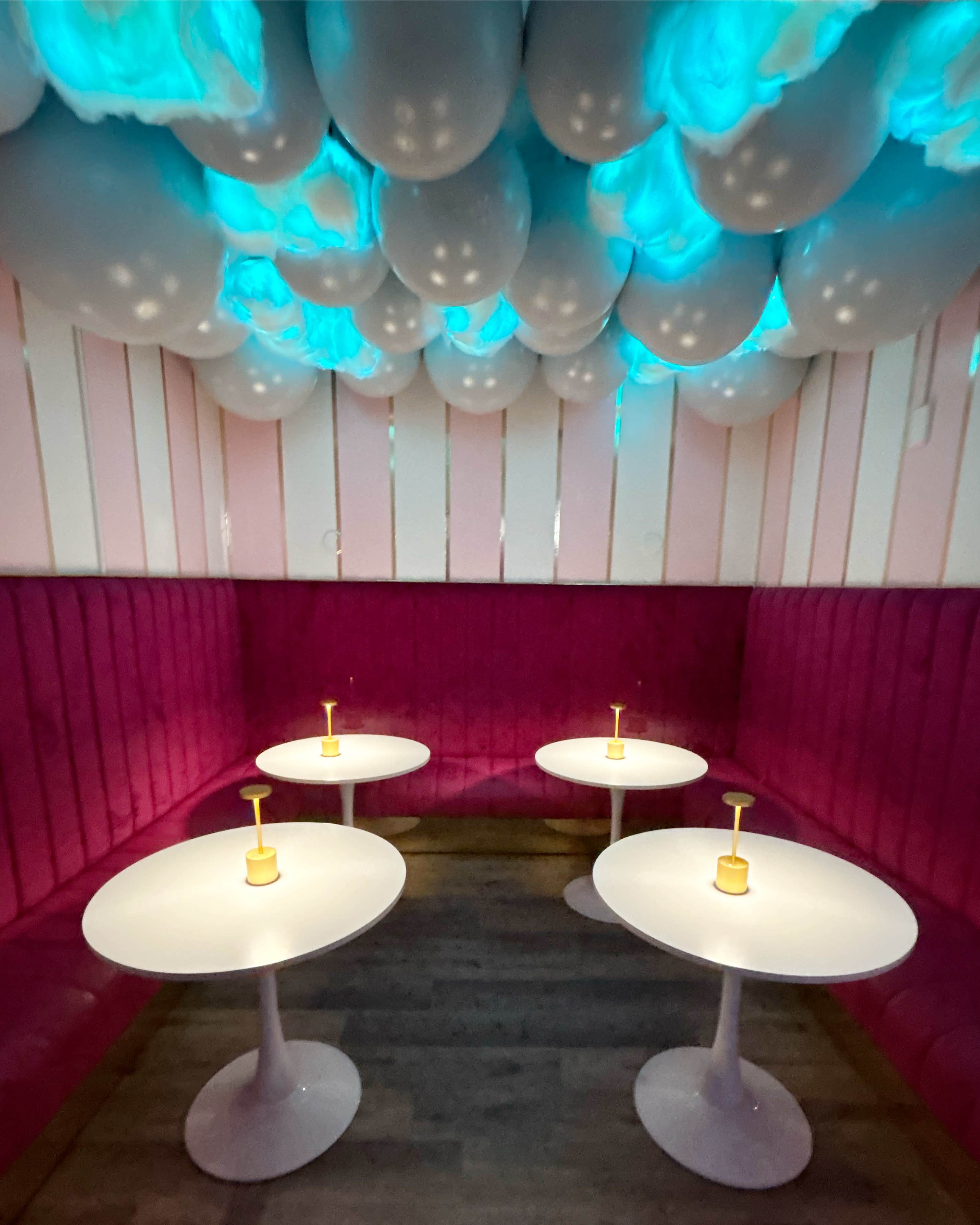A cozy lounge features four white tables with yellow drink glasses beneath a ceiling adorned with illuminated balloons.