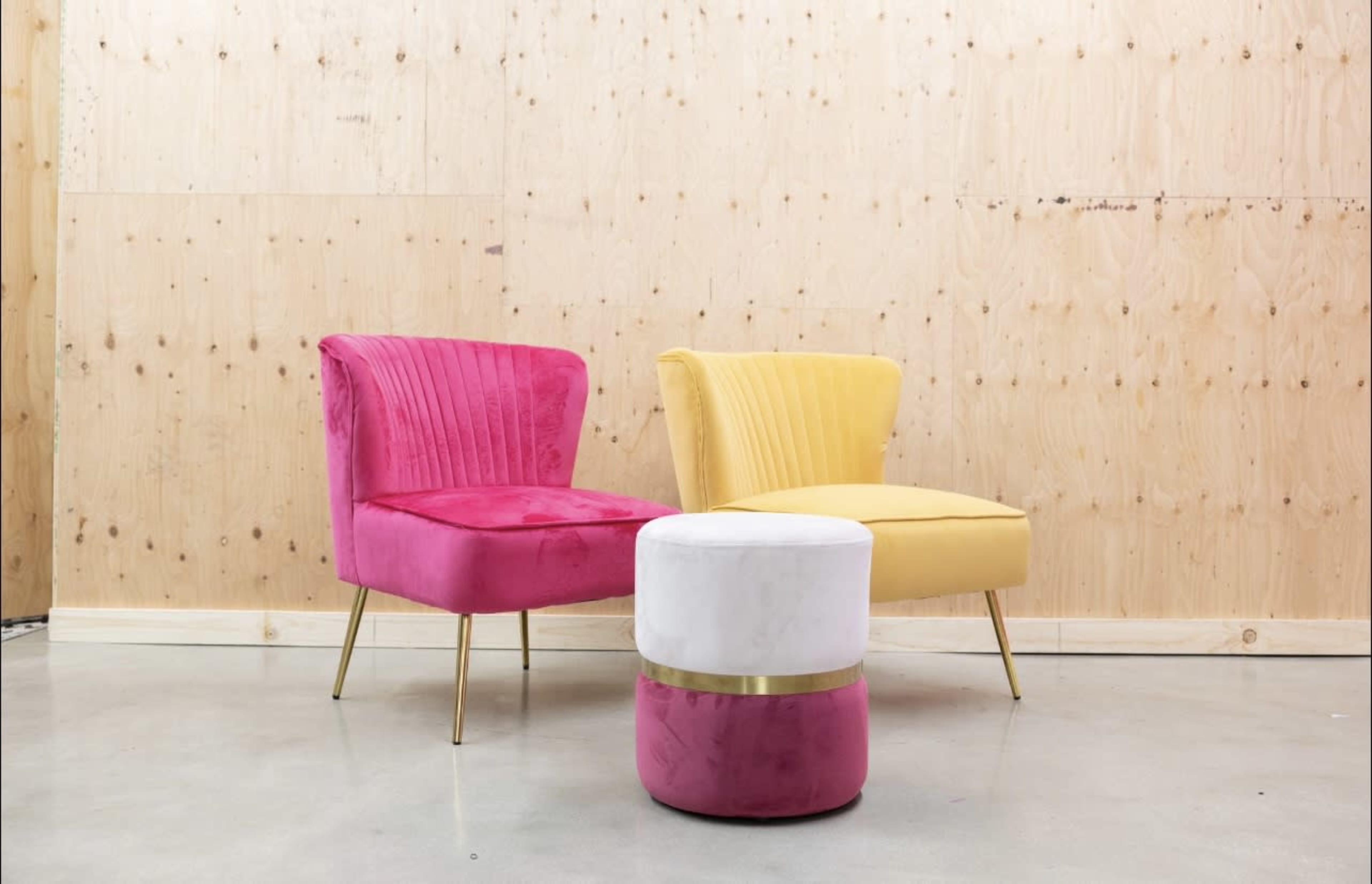 Two velvet chairs, one pink and one yellow, are placed beside a white, round ottoman with a gold band, set against a wooden wall.