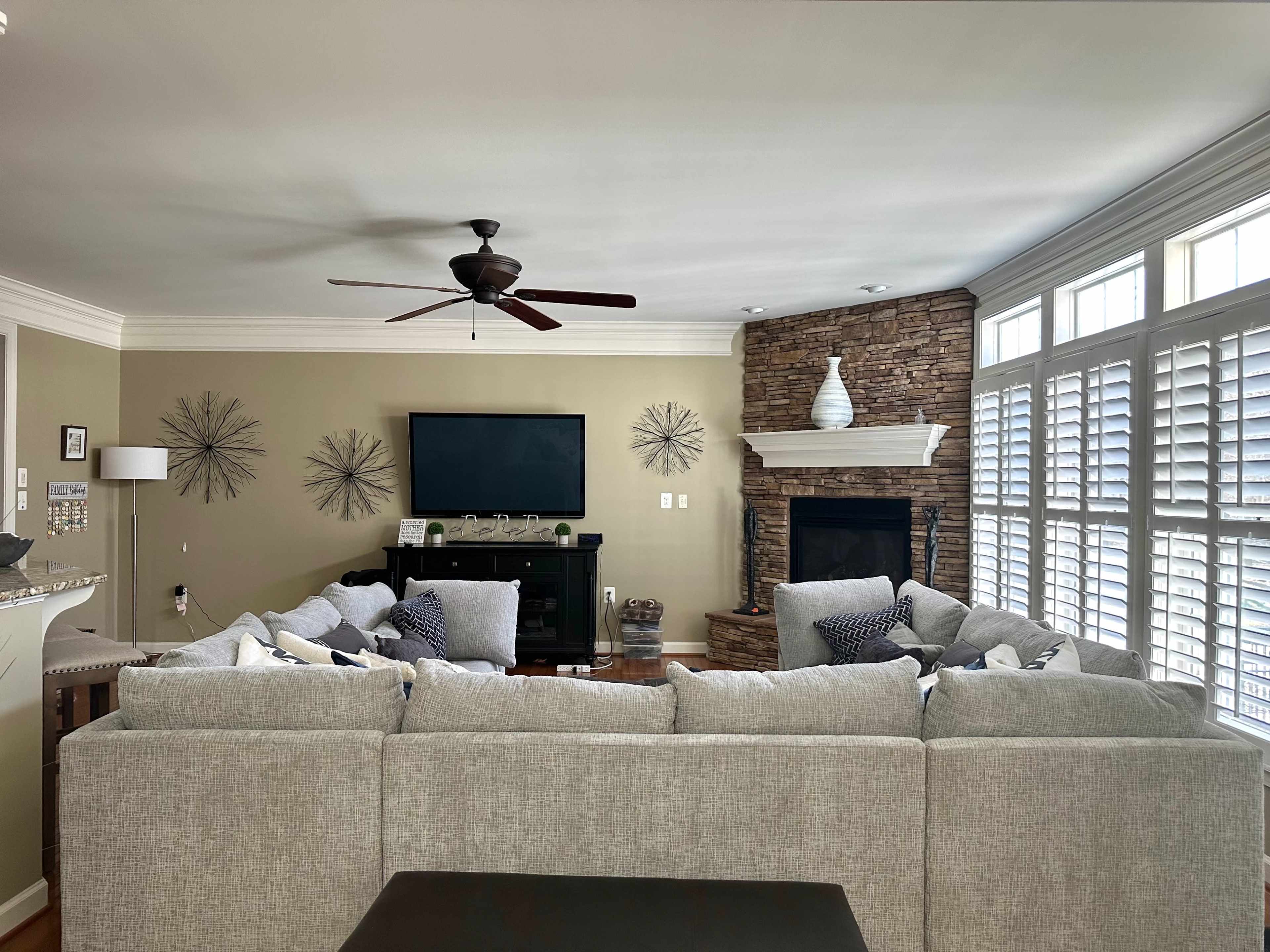 The image shows a living room featuring a large sectional sofa, a stone fireplace with a mantle, and a flat-screen television on the wall opposite large windows with plantation shutters.