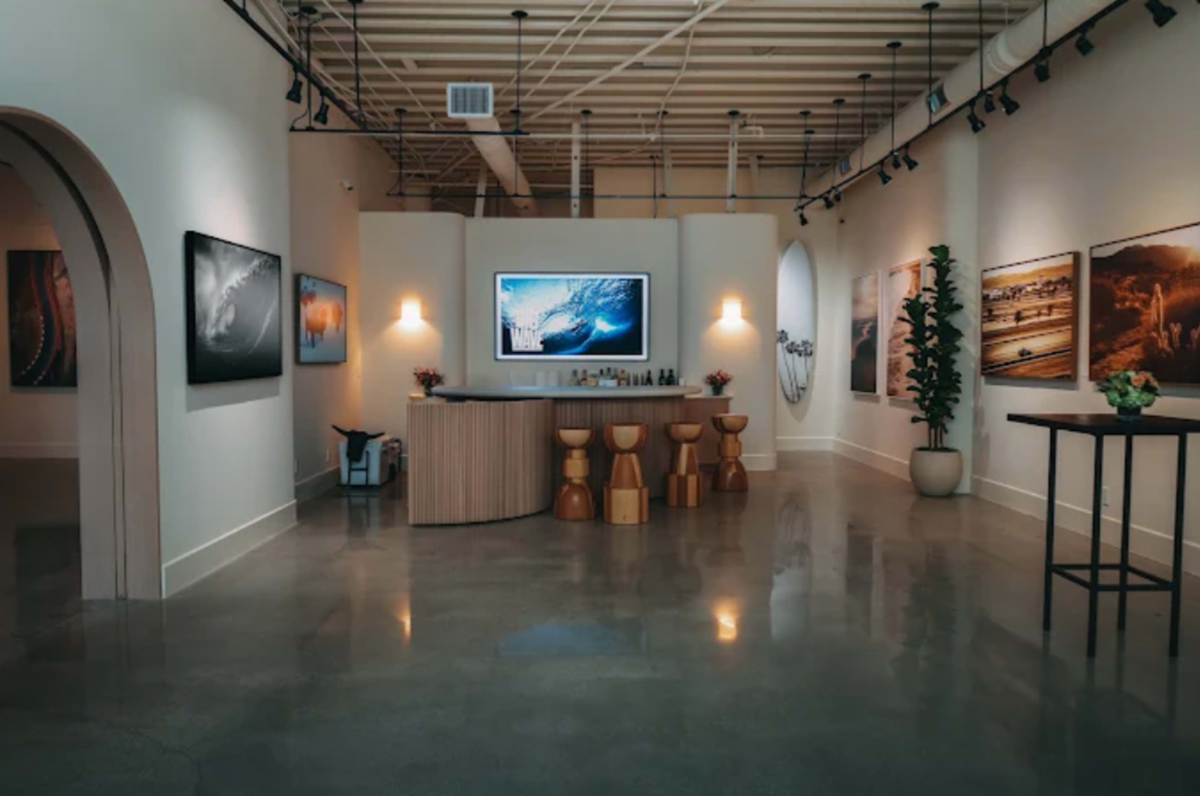 The image shows a modern art gallery with polished concrete floors, a central bar area, and several artworks displayed on the walls.