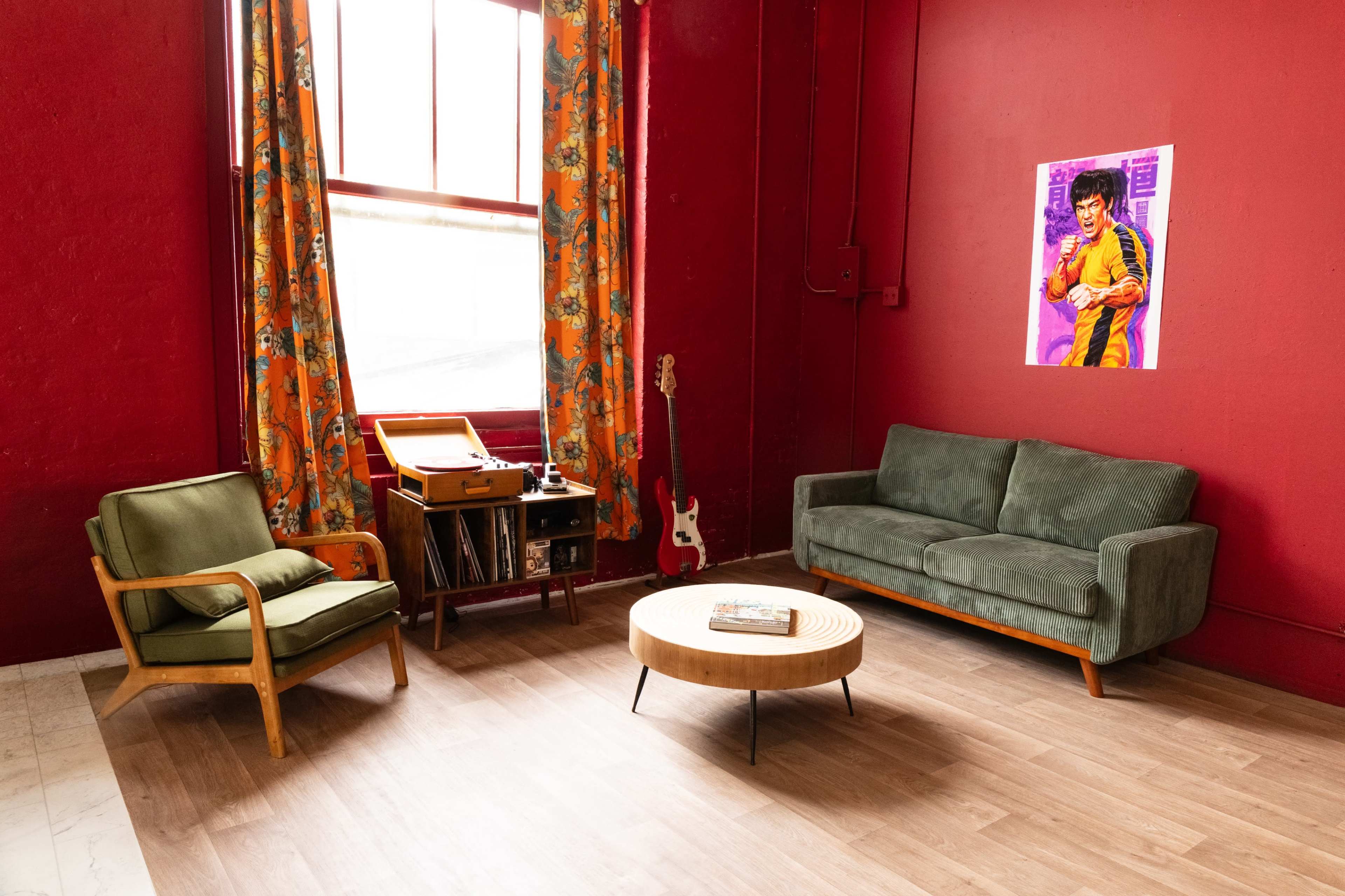 The room features red walls, a green sofa, an armchair, a round coffee table, and a vintage record player, with a colorful curtain and a framed portrait on the wall.