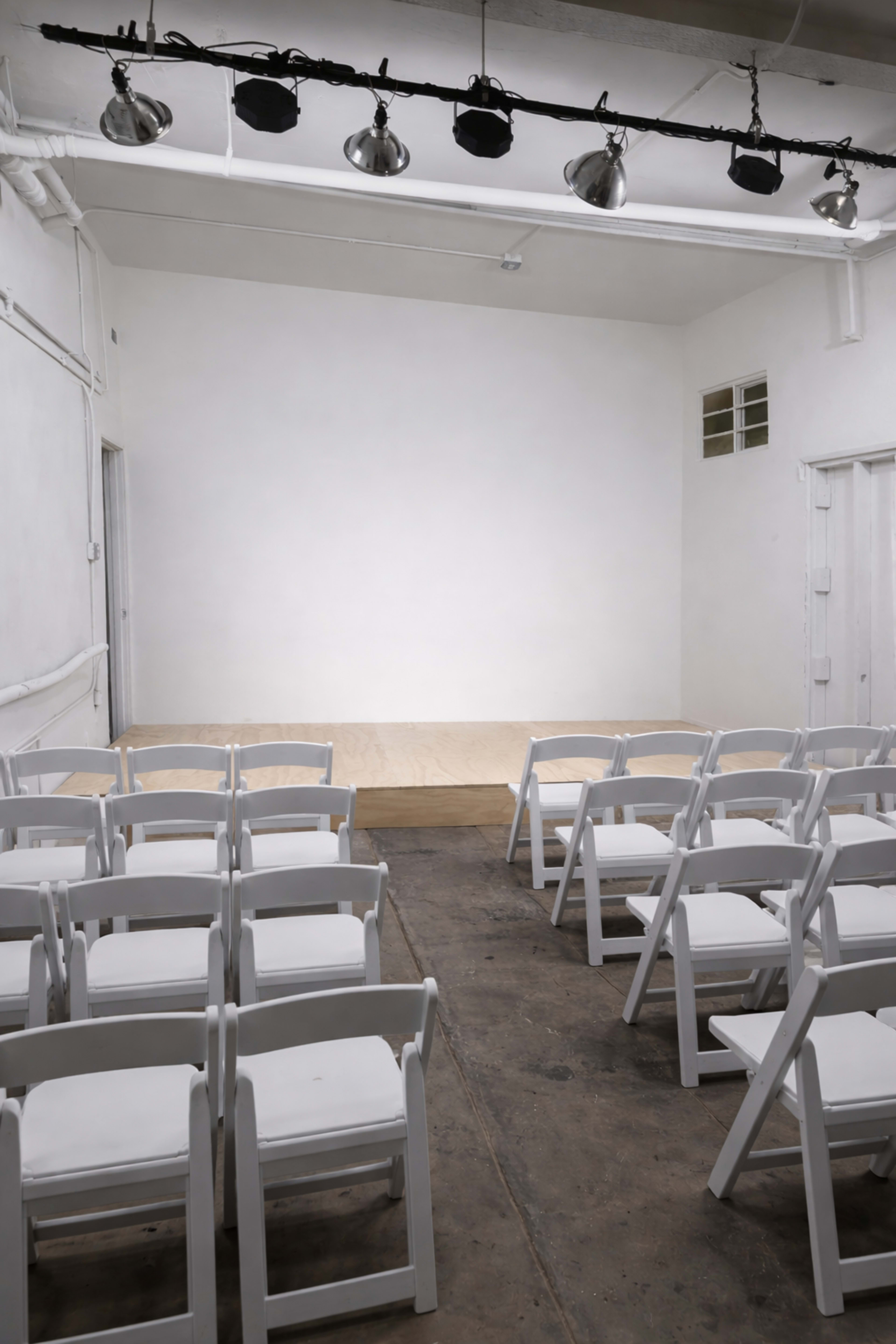 The image shows a simple, empty performance space with a wooden stage and rows of white chairs set up in front of it.