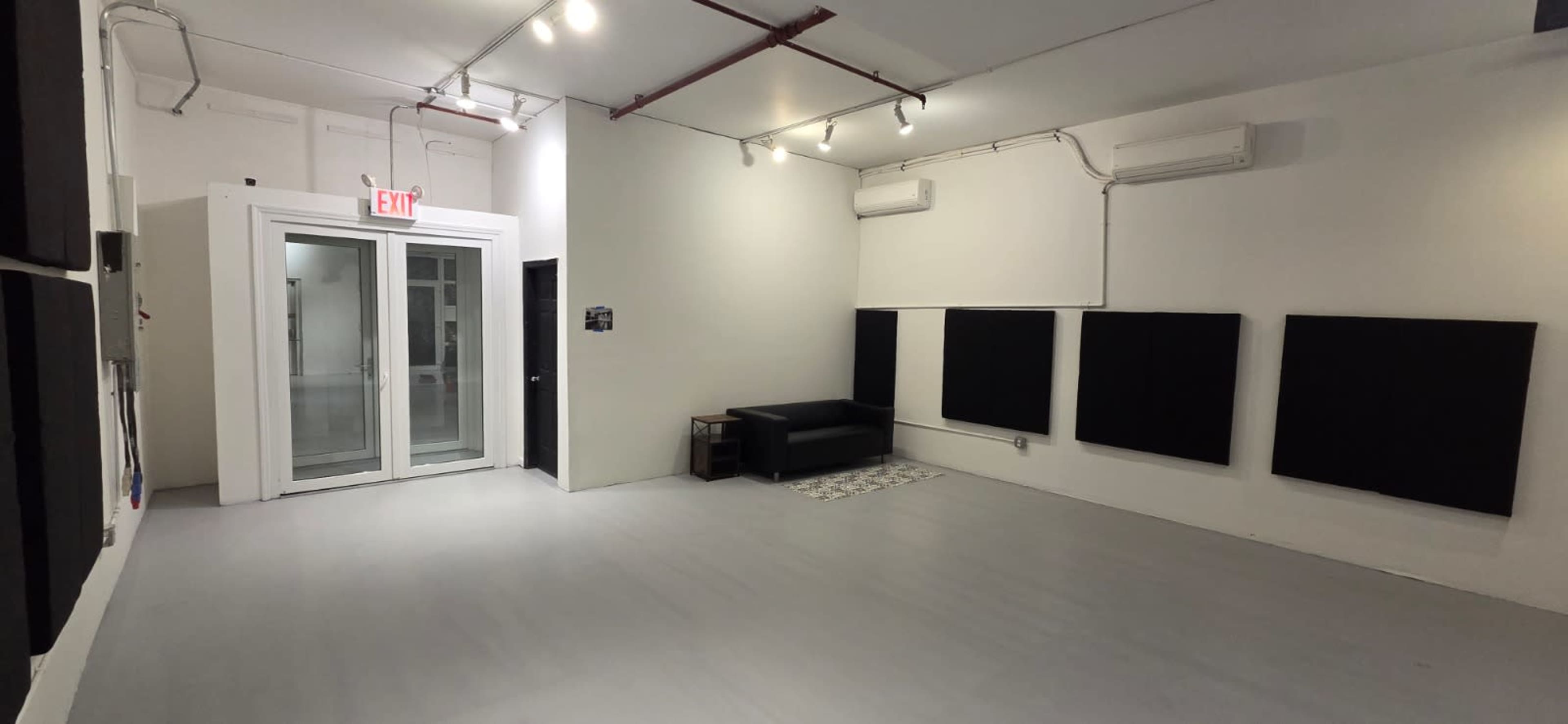 The image shows a spacious, empty room with white walls, a black sofa, and soundproofing panels on two walls.