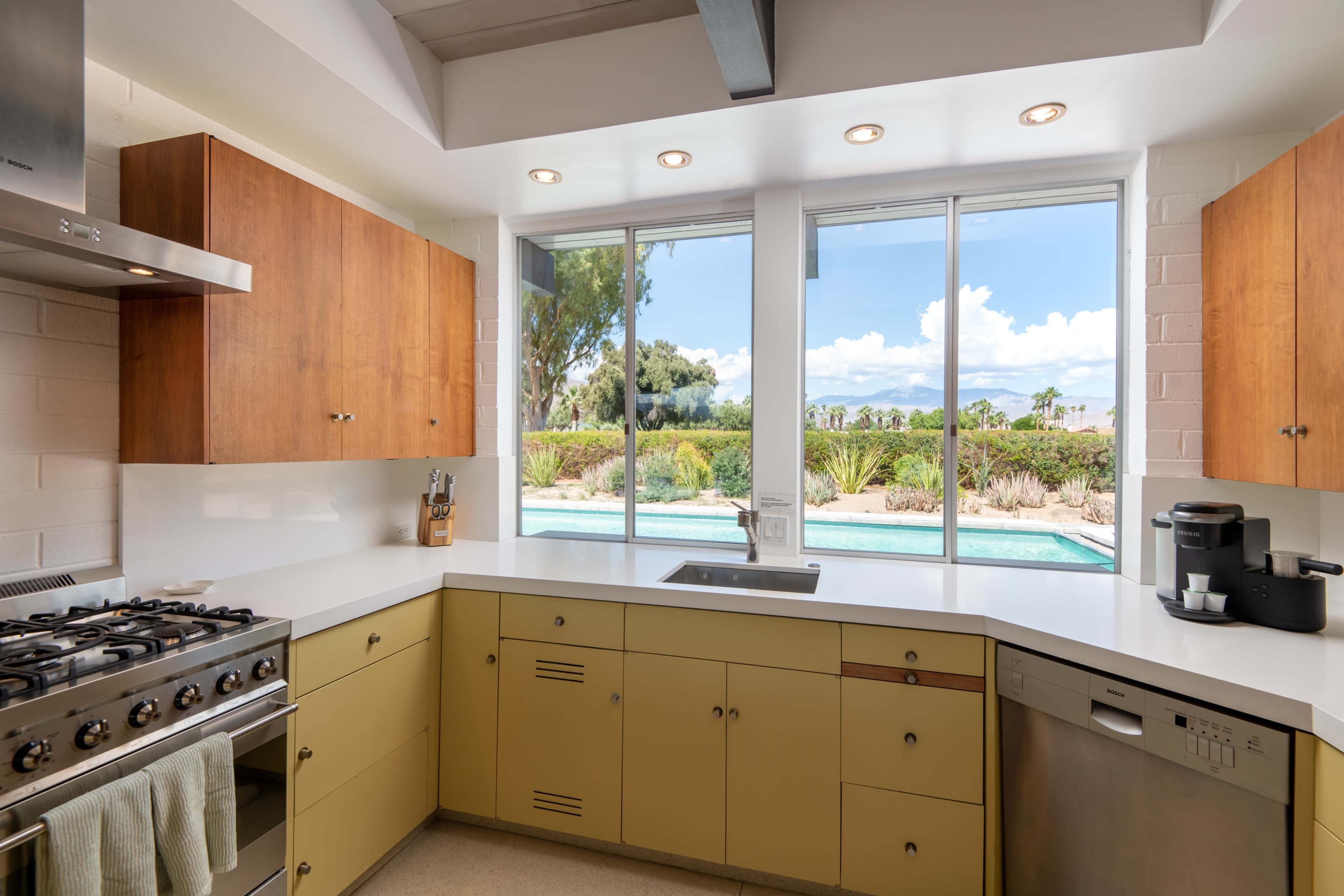 A modern kitchen features light yellow cabinets and a large window overlooking a pool and landscaped yard.