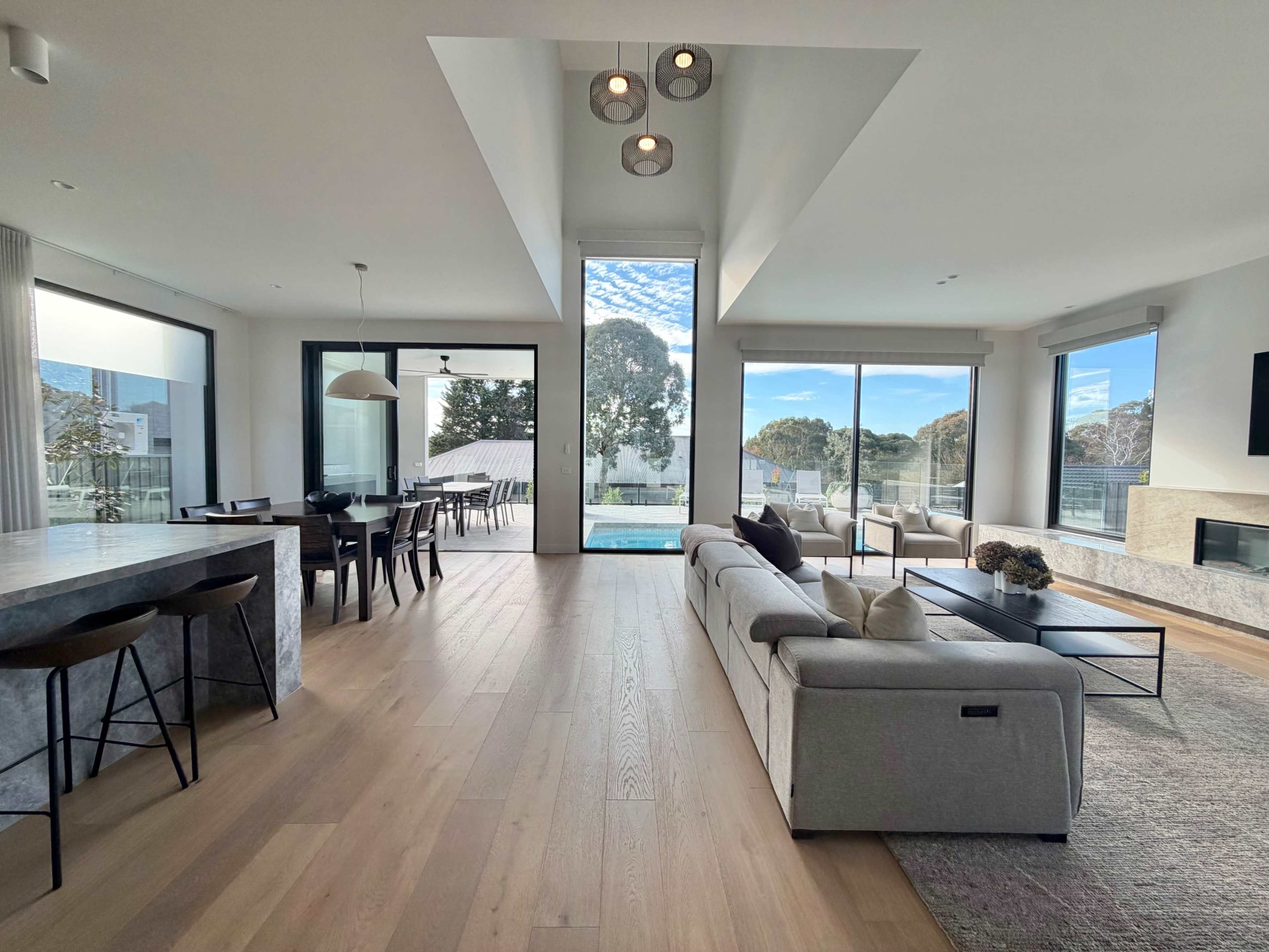 The image shows a modern, open-concept living space with a sectional sofa, dining area, and large windows overlooking a pool and outdoor scenery.