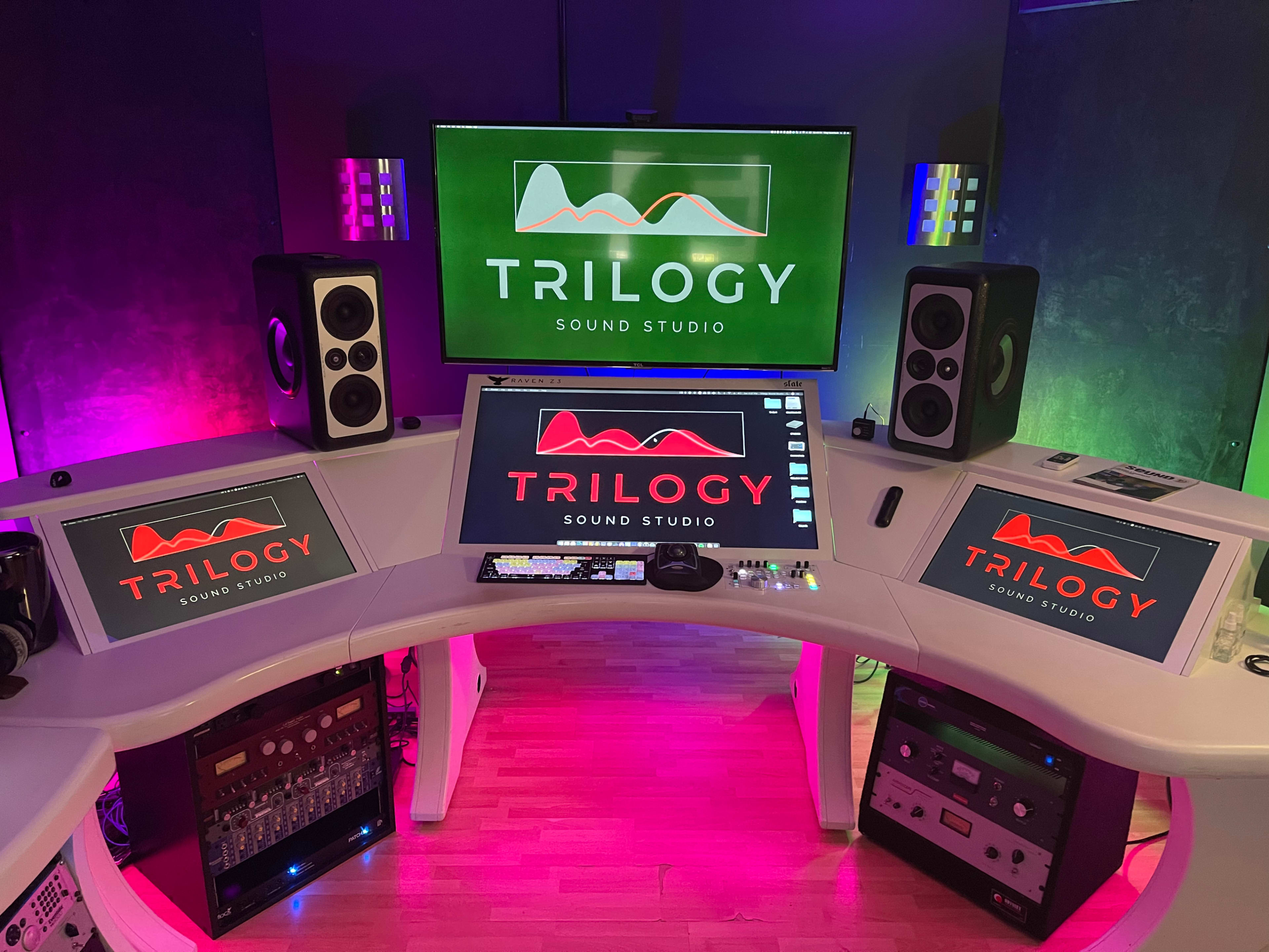 The image shows a modern sound studio setup featuring a curved desk with a computer monitor, speakers, and various audio equipment against a backdrop of illuminated green and purple walls.