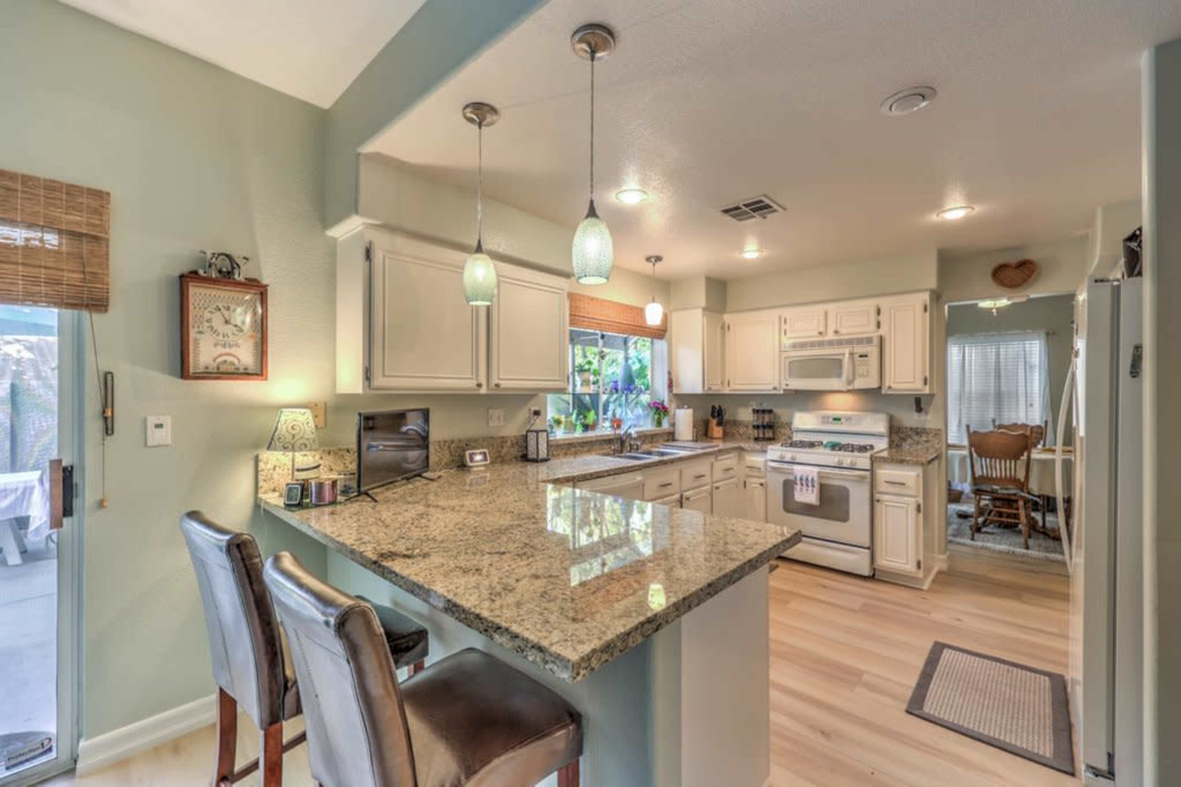 A modern kitchen features a granite island with bar seating, white cabinetry, and appliances, illuminated by pendant lights.