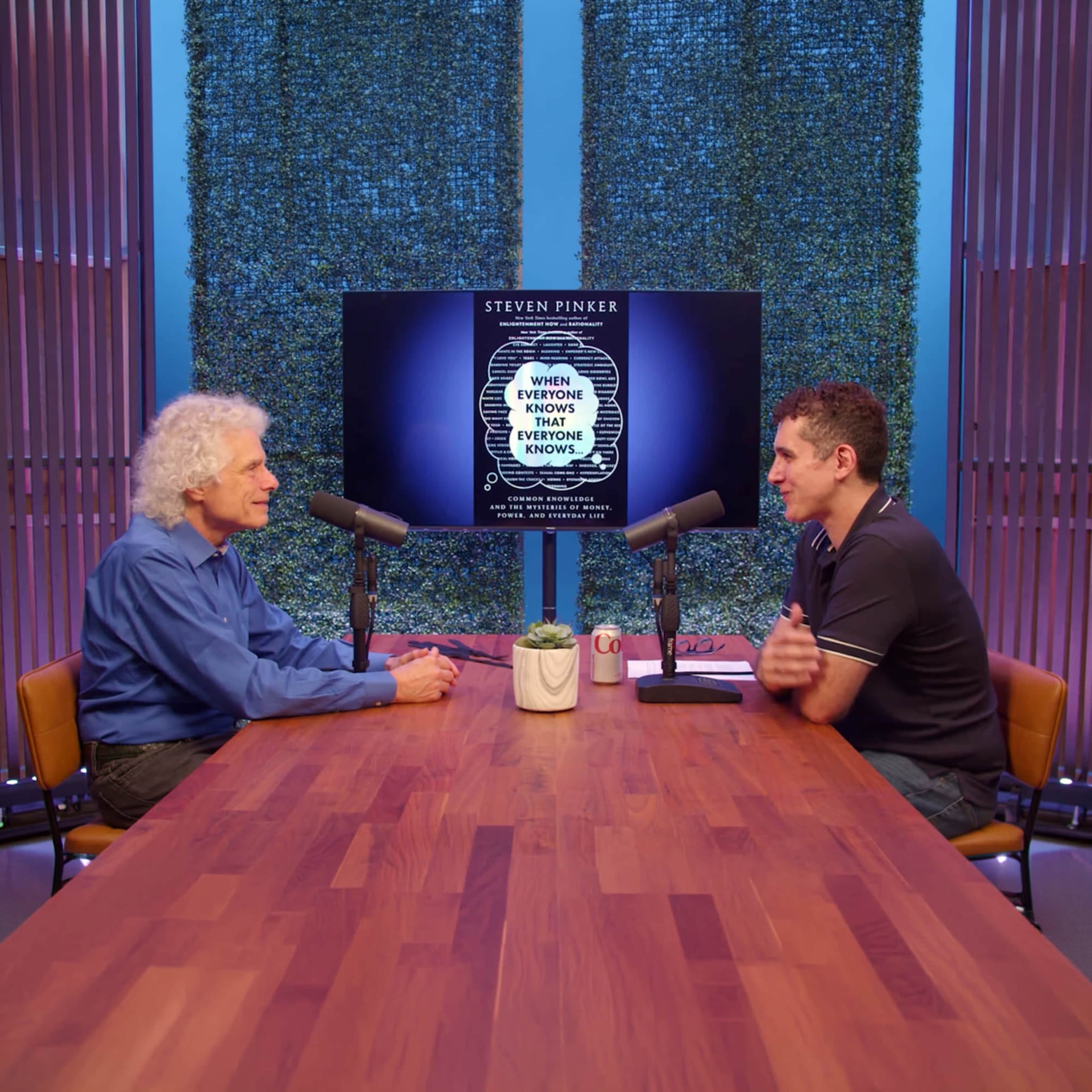 Two men are seated at a table with microphones, engaged in a conversation, while a screen displaying the title of a book by Steven Pinker is in the background.