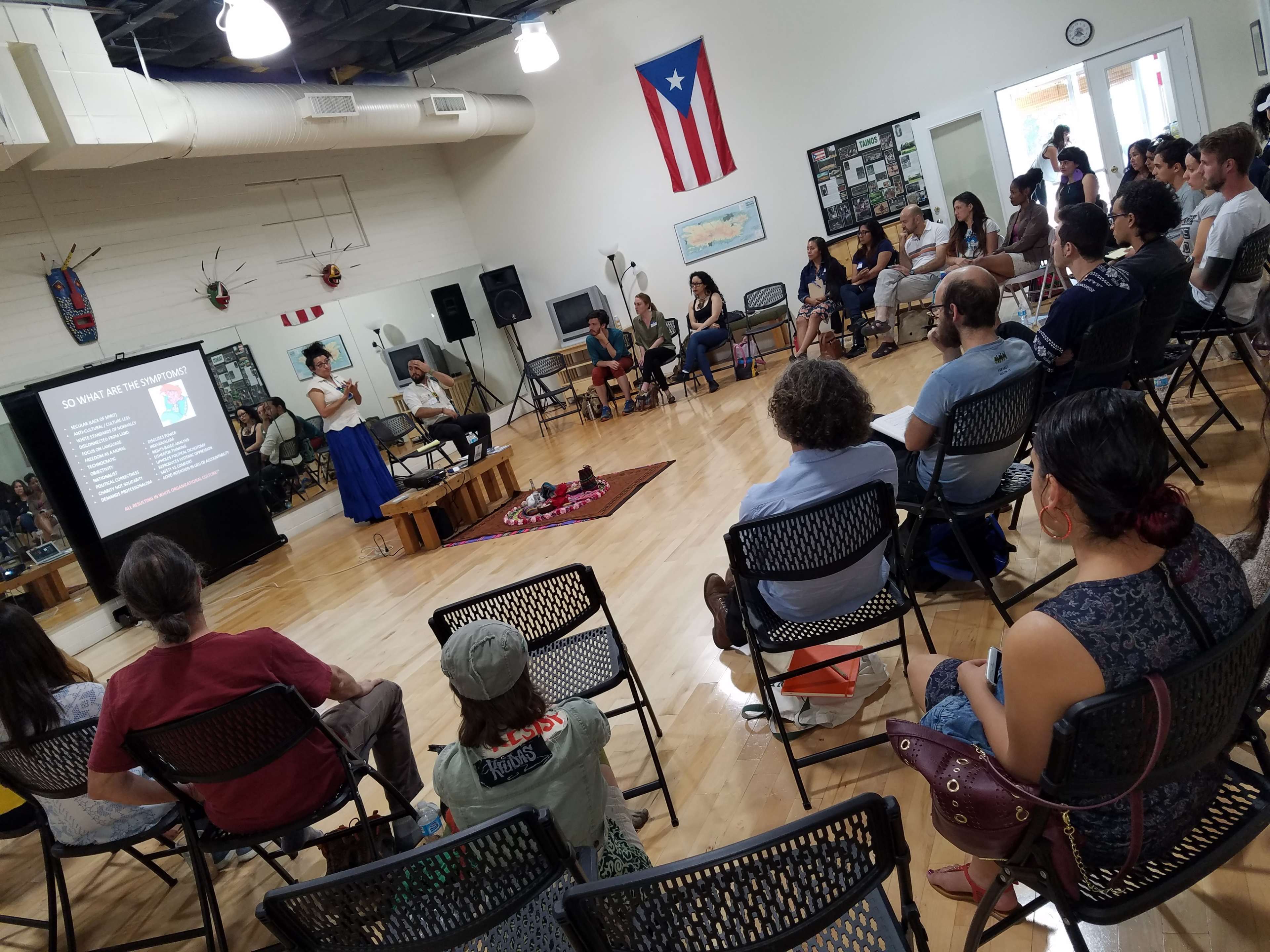 A speaker presents to an audience seated in a community center, with a projector screen displaying information in the background.