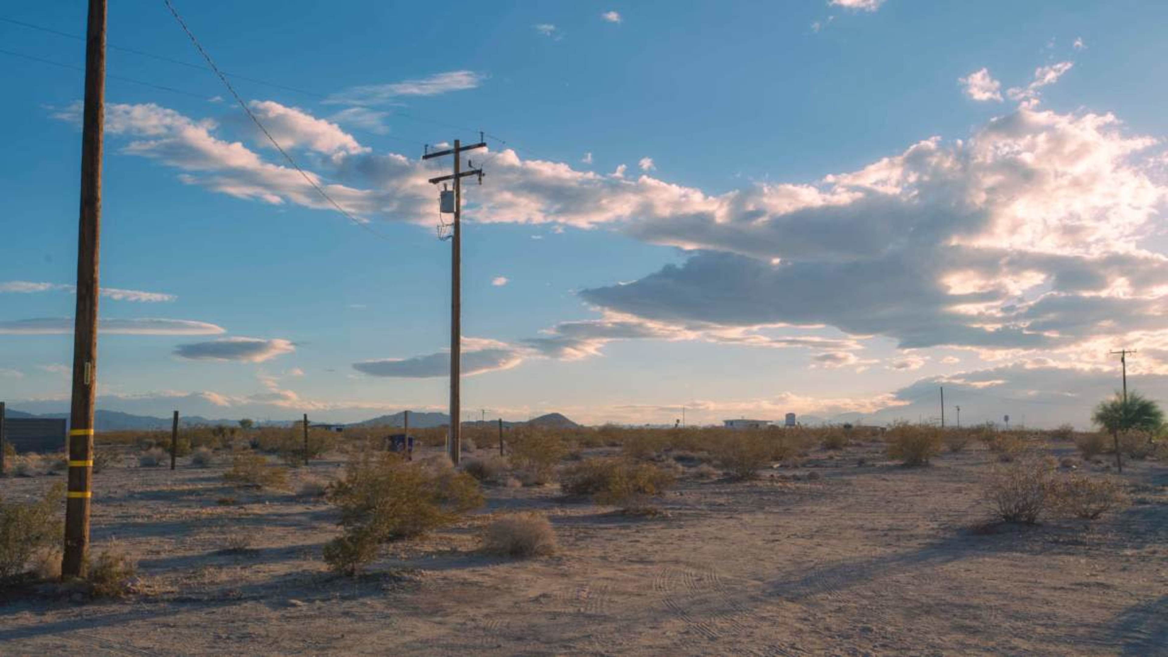 A barren landscape features telephone poles amidst sparse vegetation under a blue sky with scattered clouds.