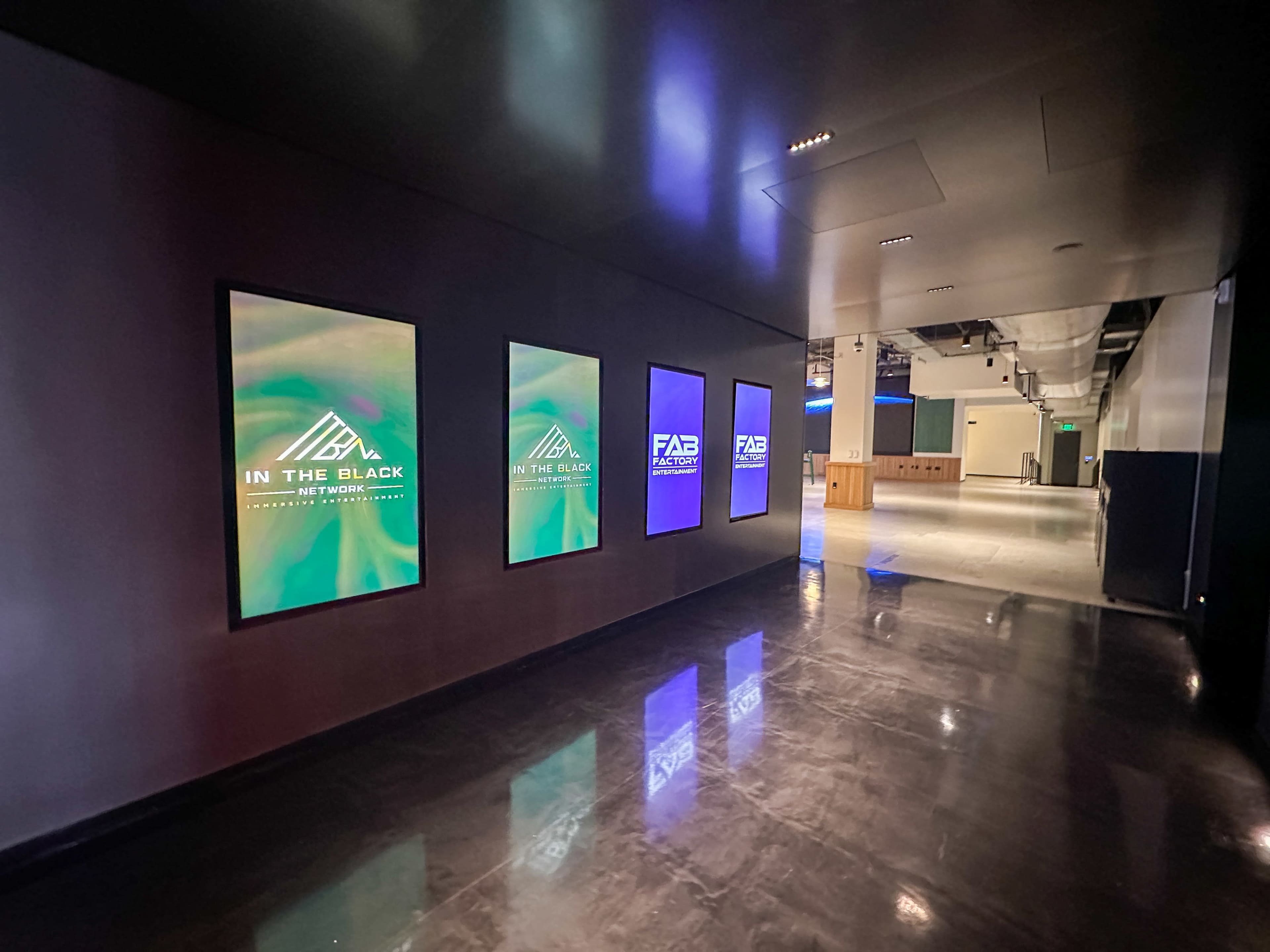 A dimly lit corridor displays several illuminated signs on the walls promoting different events or networks.