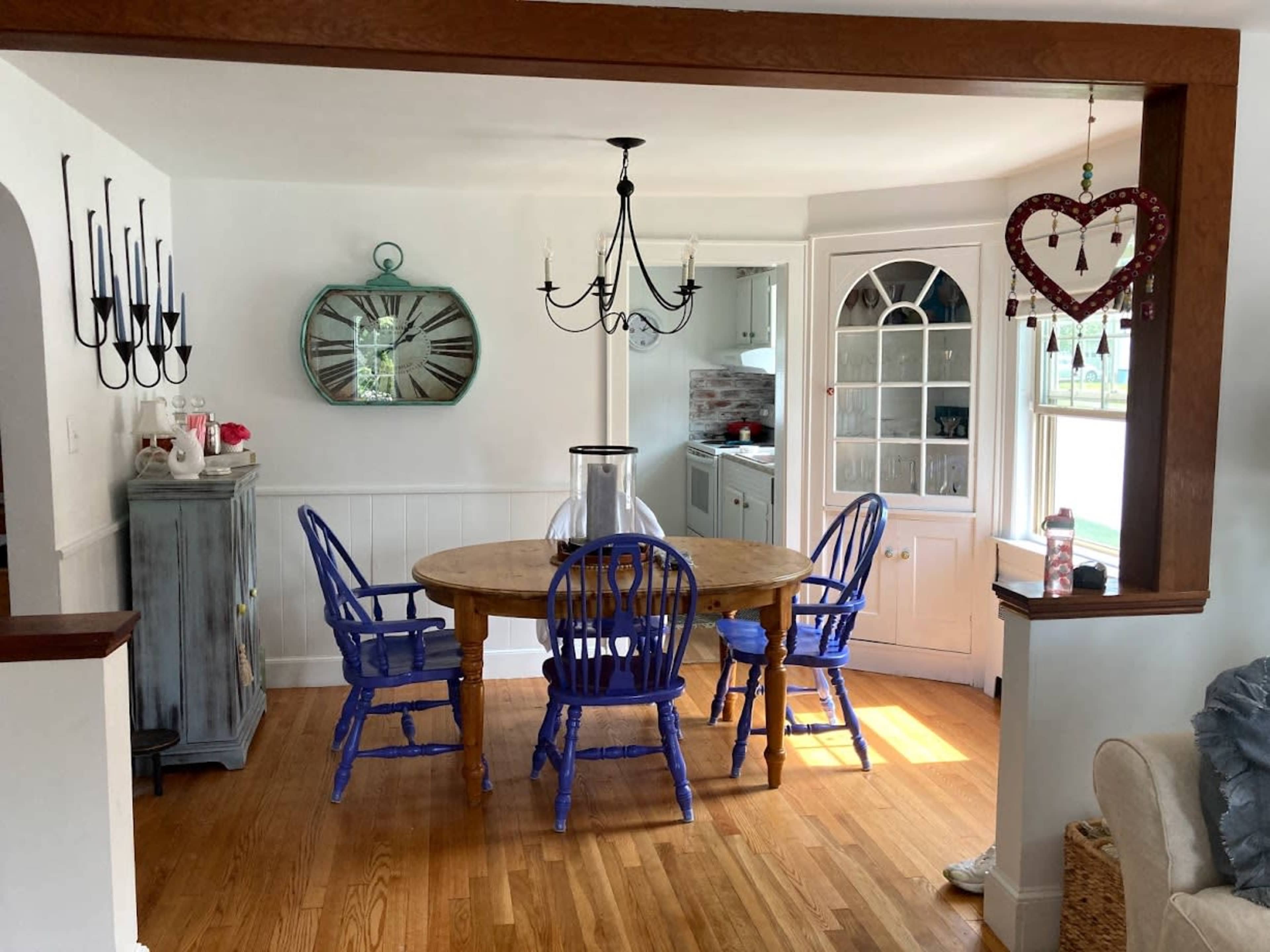 A dining area features a round wooden table surrounded by blue chairs, with a chandelier above and a large mirror on the wall.