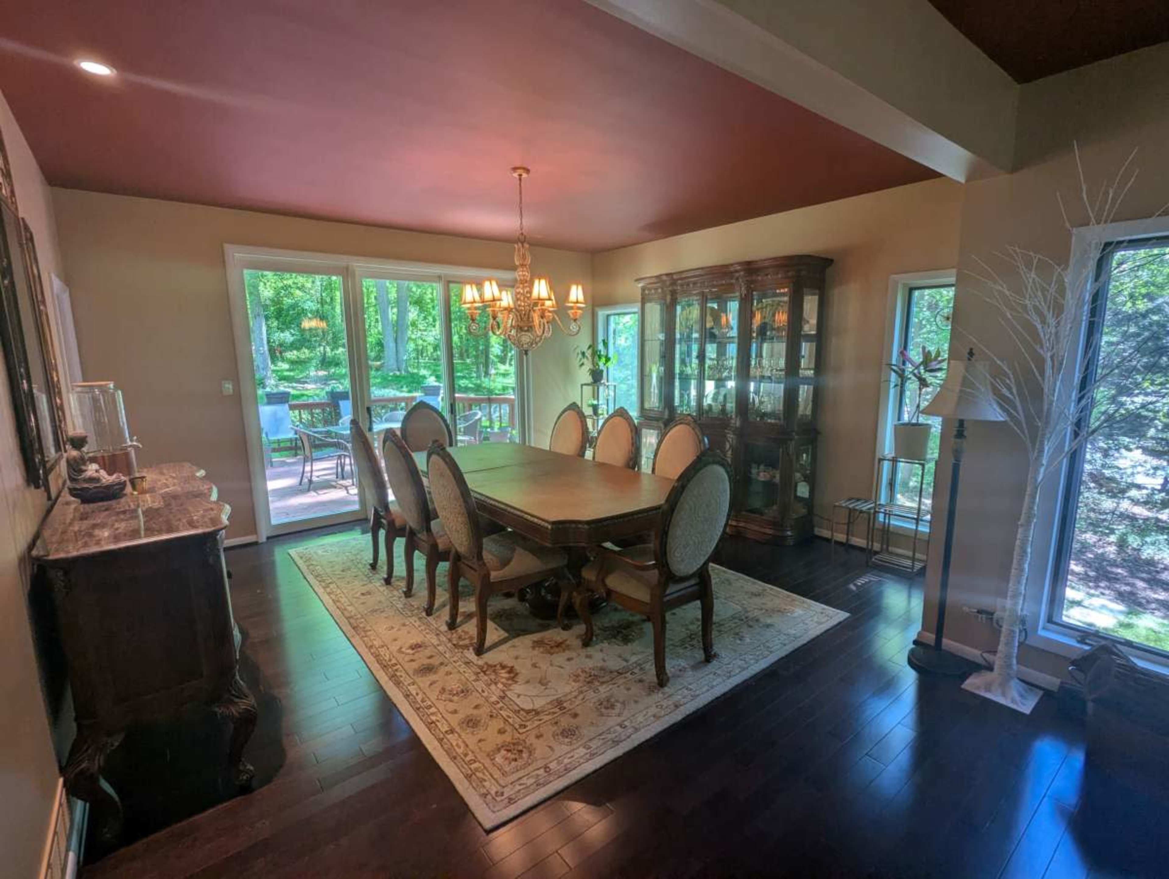 The image shows a dining room with a large wooden table surrounded by chairs, a chandelier overhead, and large windows offering a view of greenery outside.