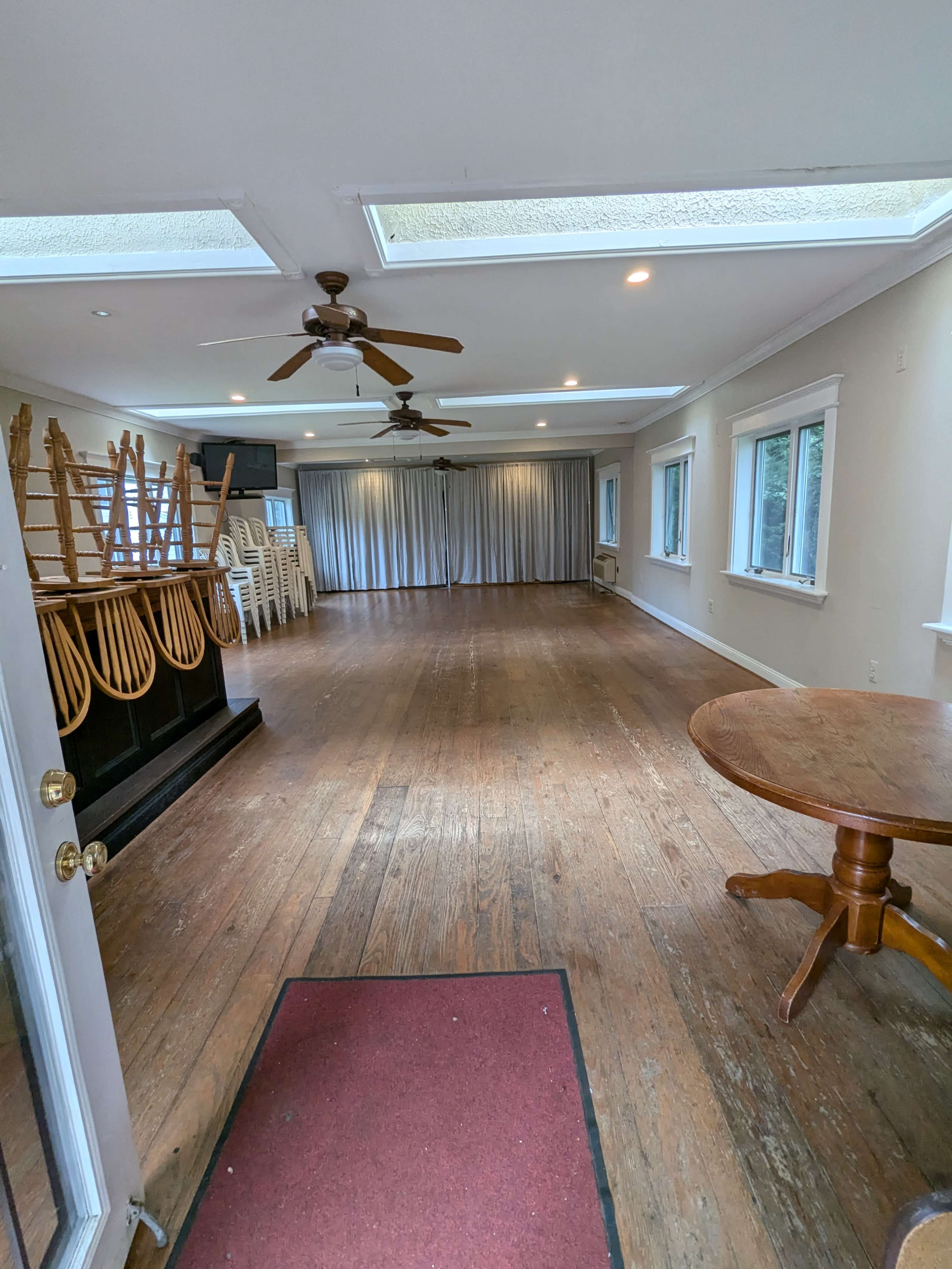 A spacious, empty room with wooden floors, chairs stacked against one wall, and large windows along the sides.