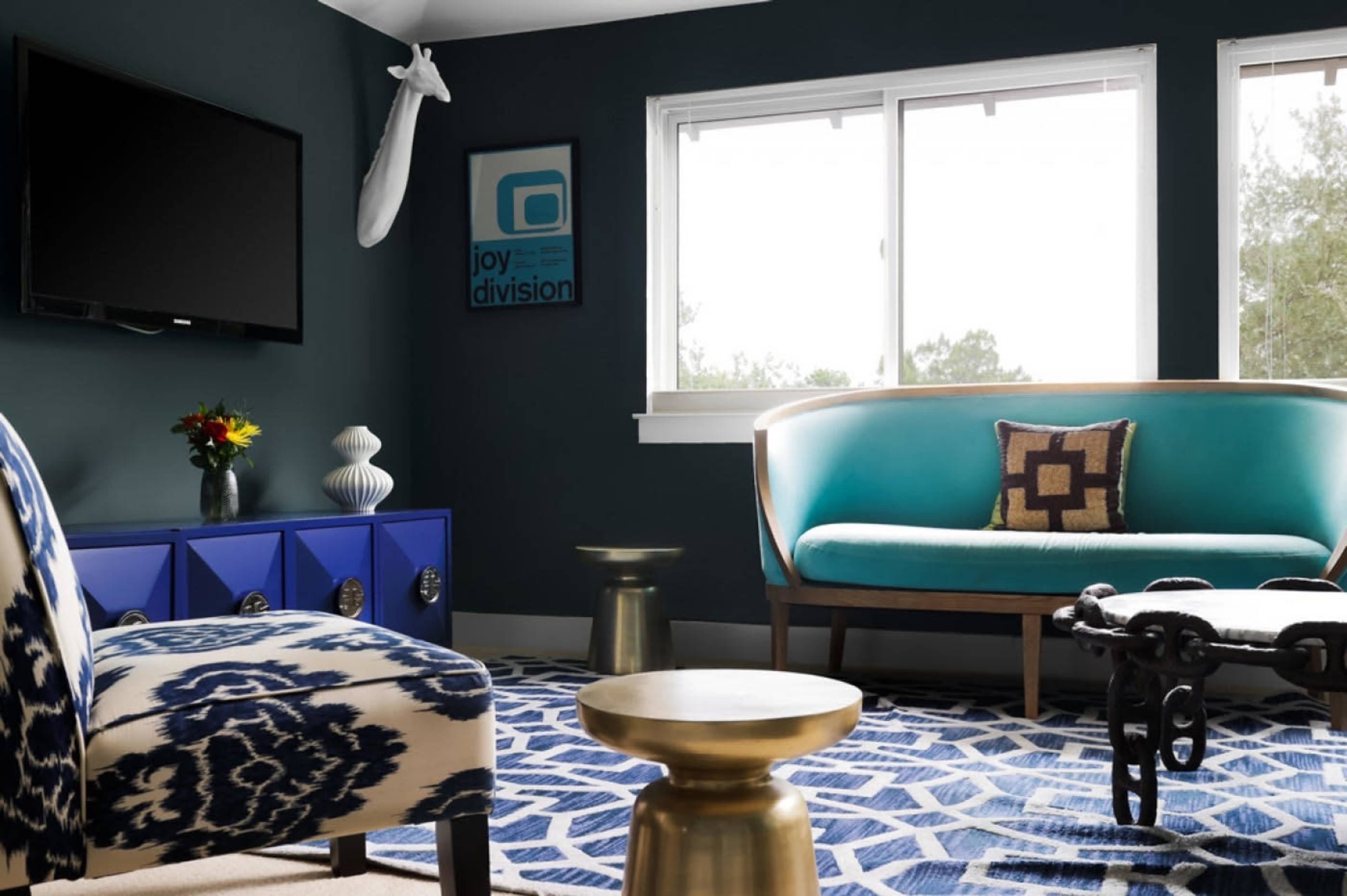 The image shows a modern living room featuring a blue and white patterned rug, a teal sofa, a patterned armchair, and a dark blue accent wall.