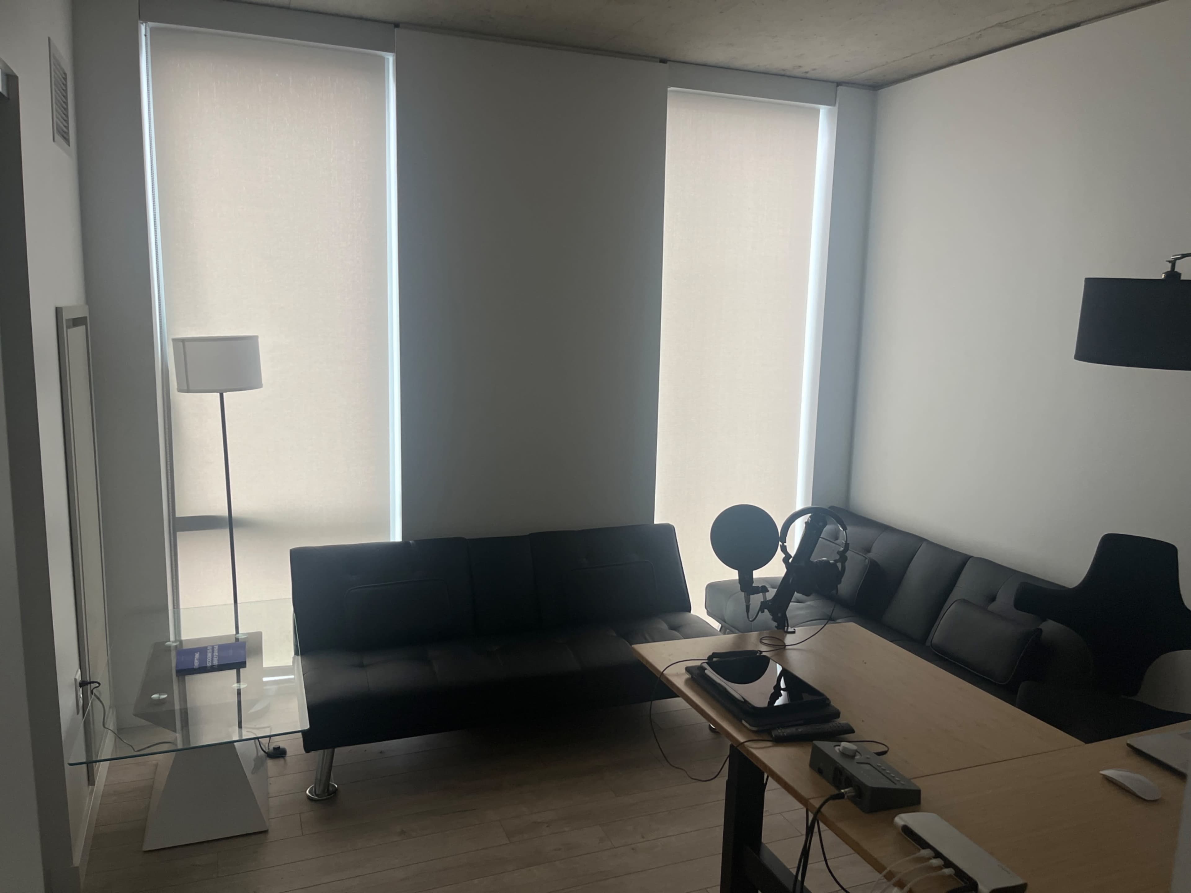 A minimalist room with a black sofa, a glass table, a microphone on the table, and large window shades allowing diffused light to enter.