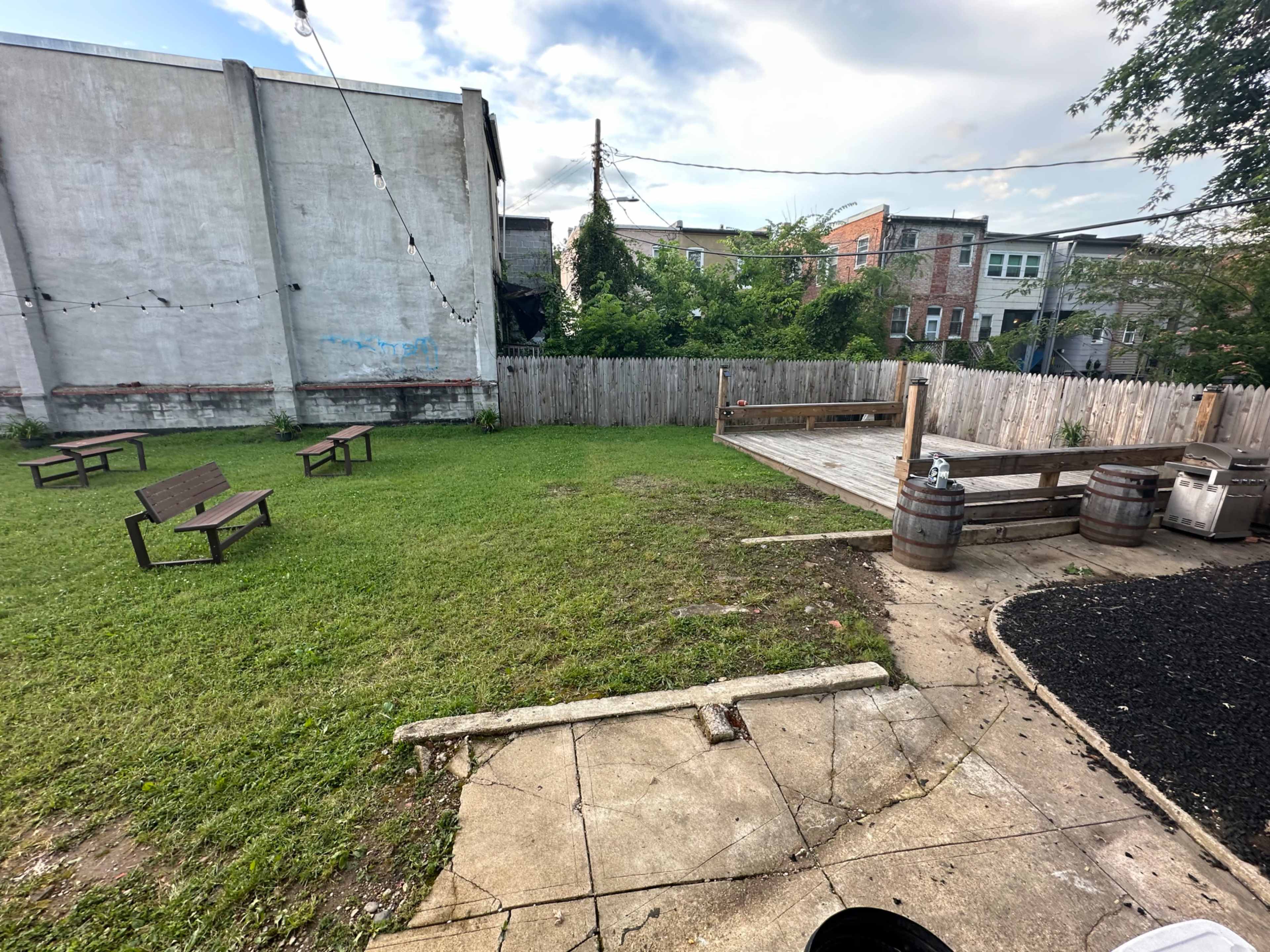 The image shows a grassy area with two wooden benches, a wooden deck, and a fenced perimeter, set against a backdrop of buildings.