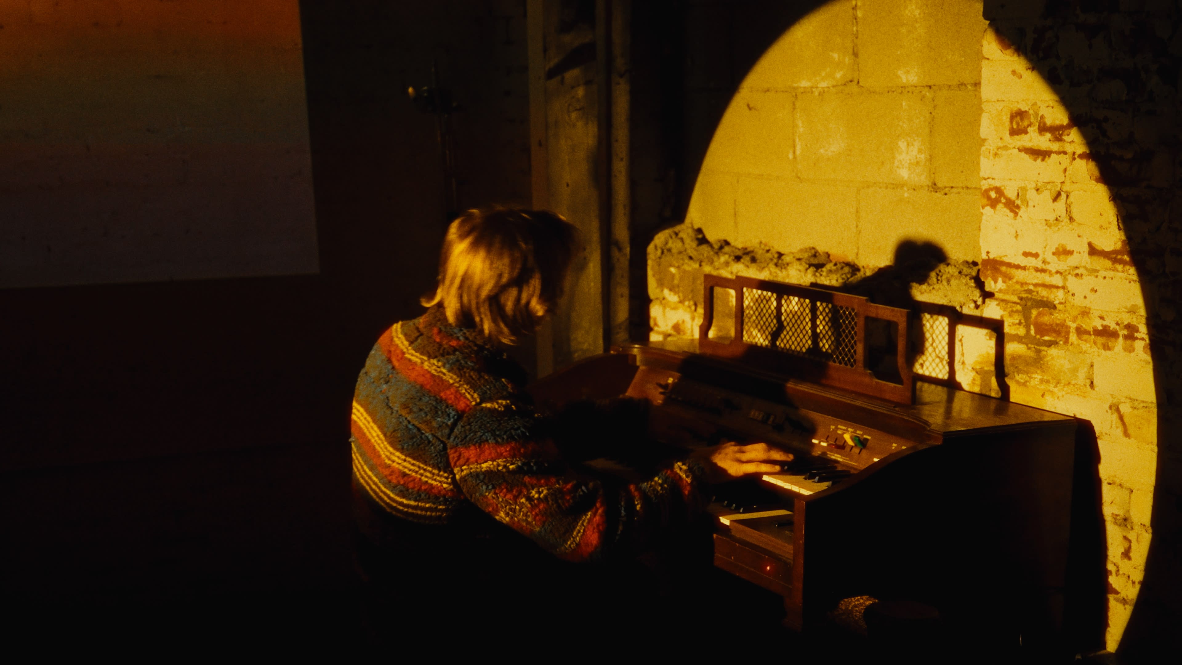 A person in a colorful sweater plays an organ in a dimly lit room with brick walls.