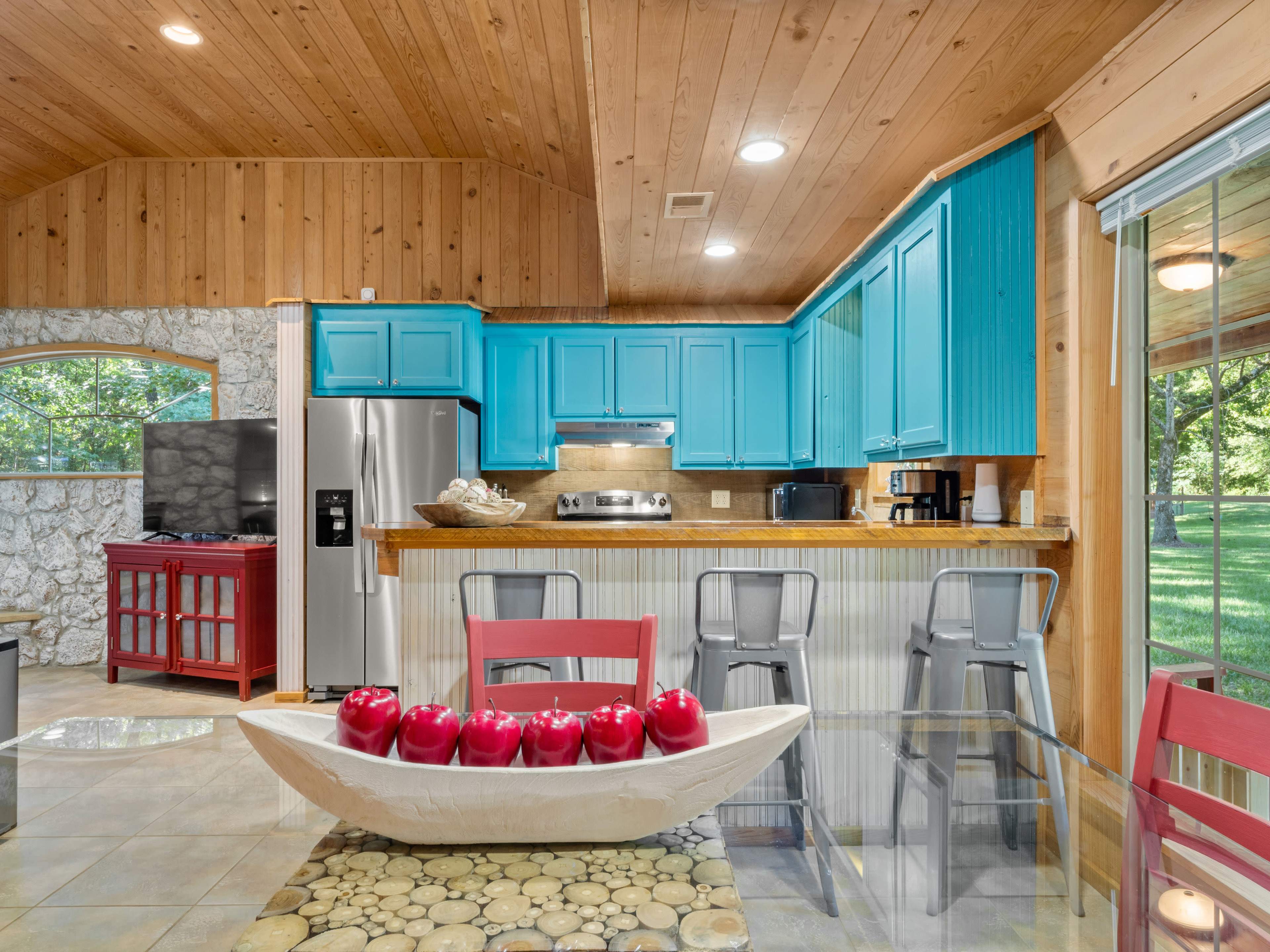 A kitchen with turquoise cabinets, stainless steel appliances, and a dining table featuring a bowl of red apples at the center.
