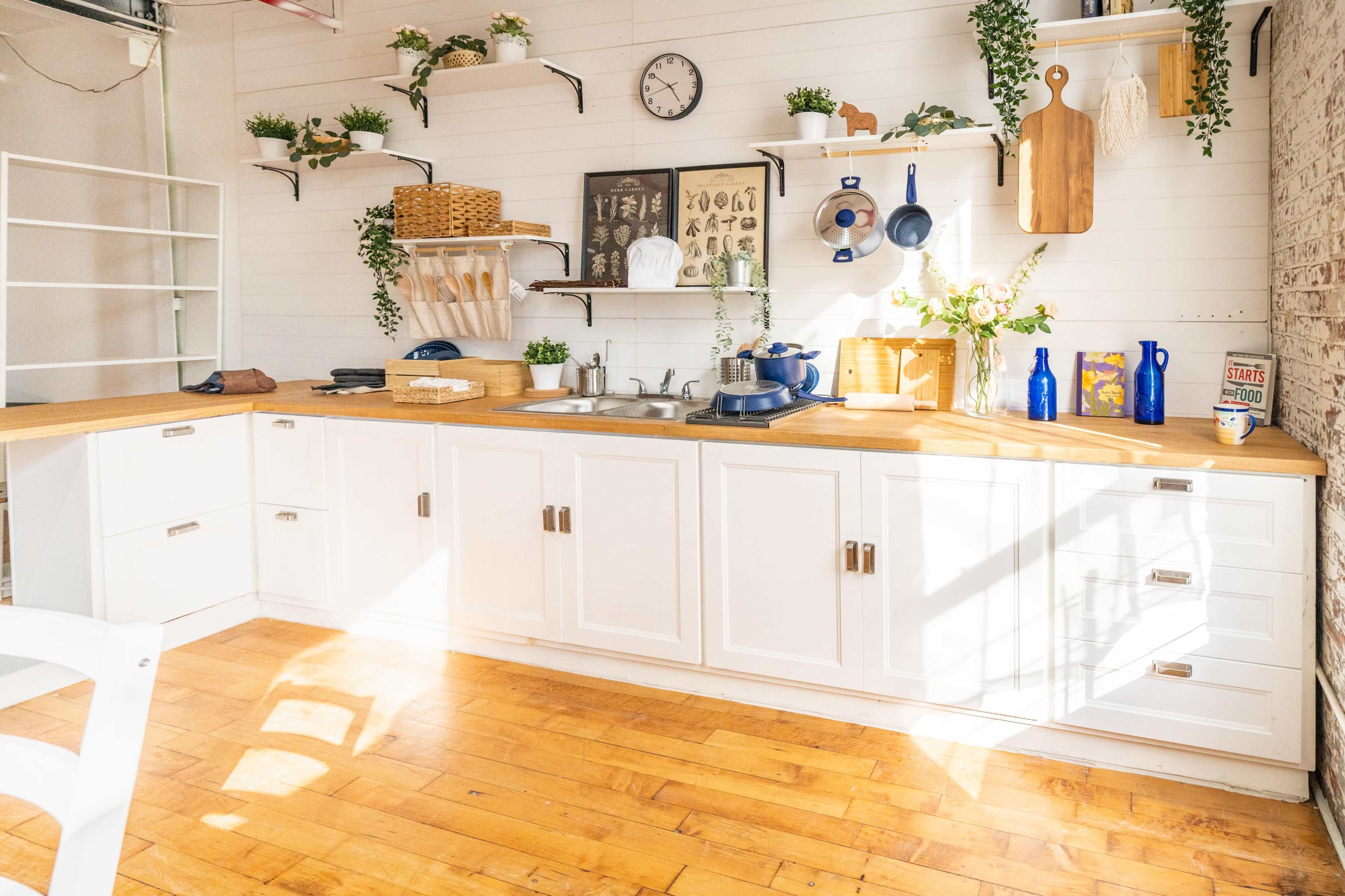A bright kitchen with white cabinets, a wooden countertop, and various decorations, including plants and kitchenware neatly arranged on shelves.