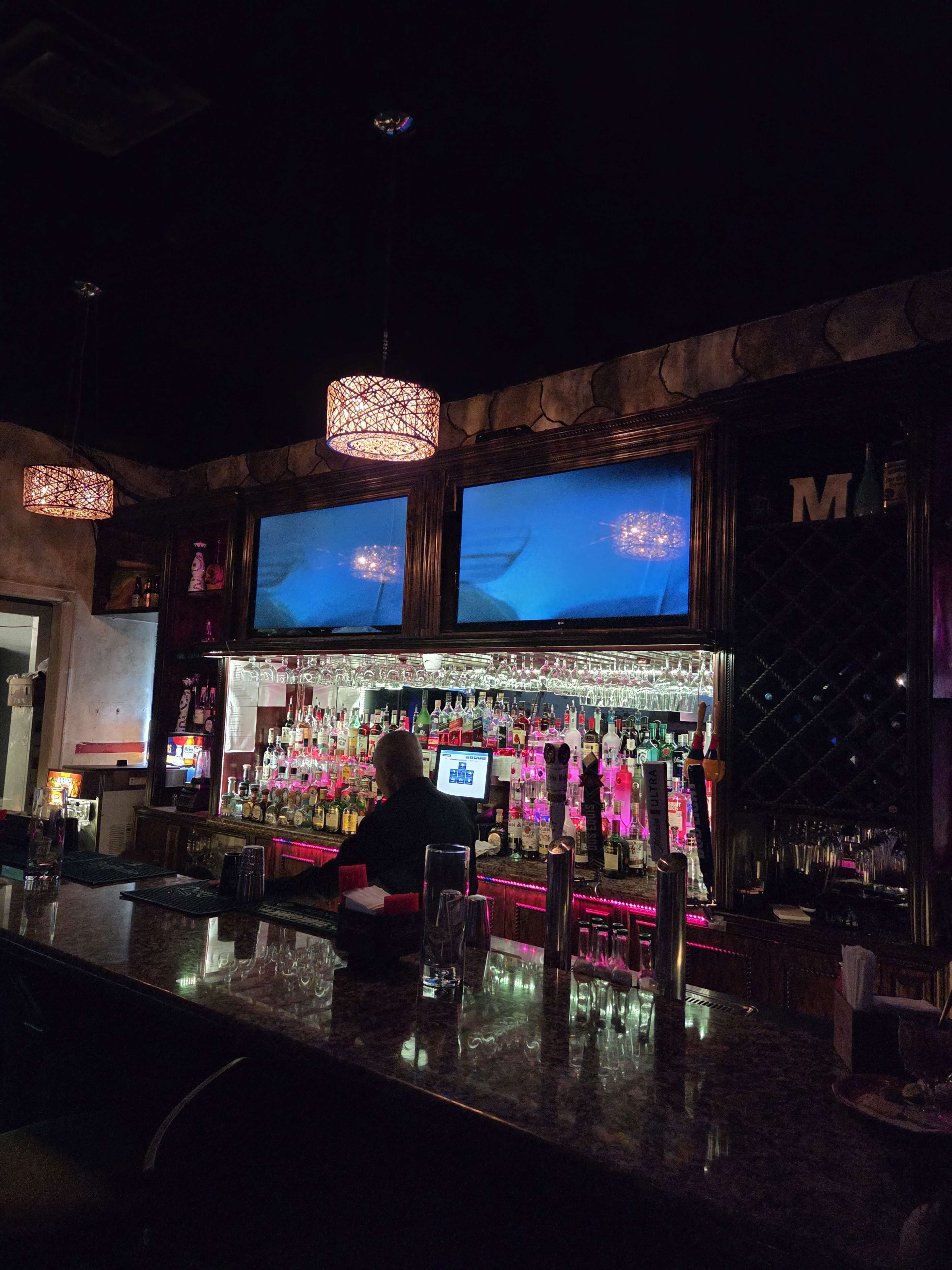 A bartender stands behind a well-stocked bar lined with colorful liquor bottles and illuminated by decorative pendant lights.
