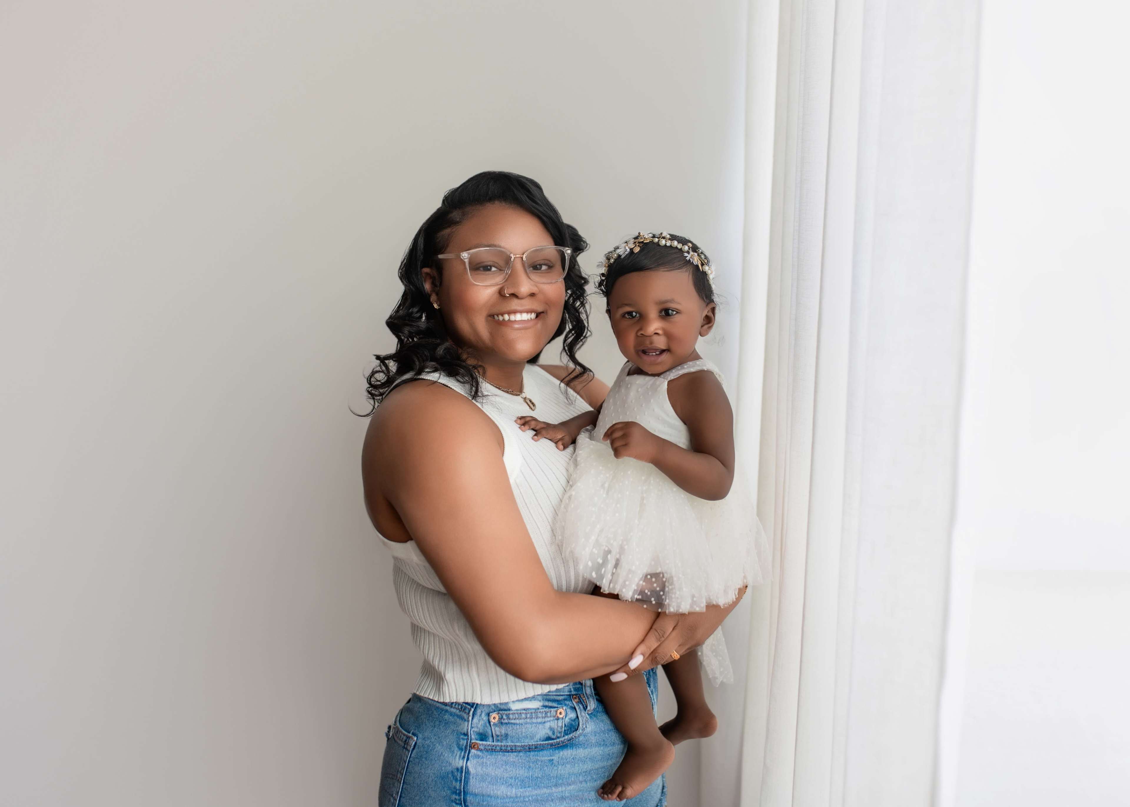 A woman with glasses holds a smiling toddler in a white dress against a light backdrop.