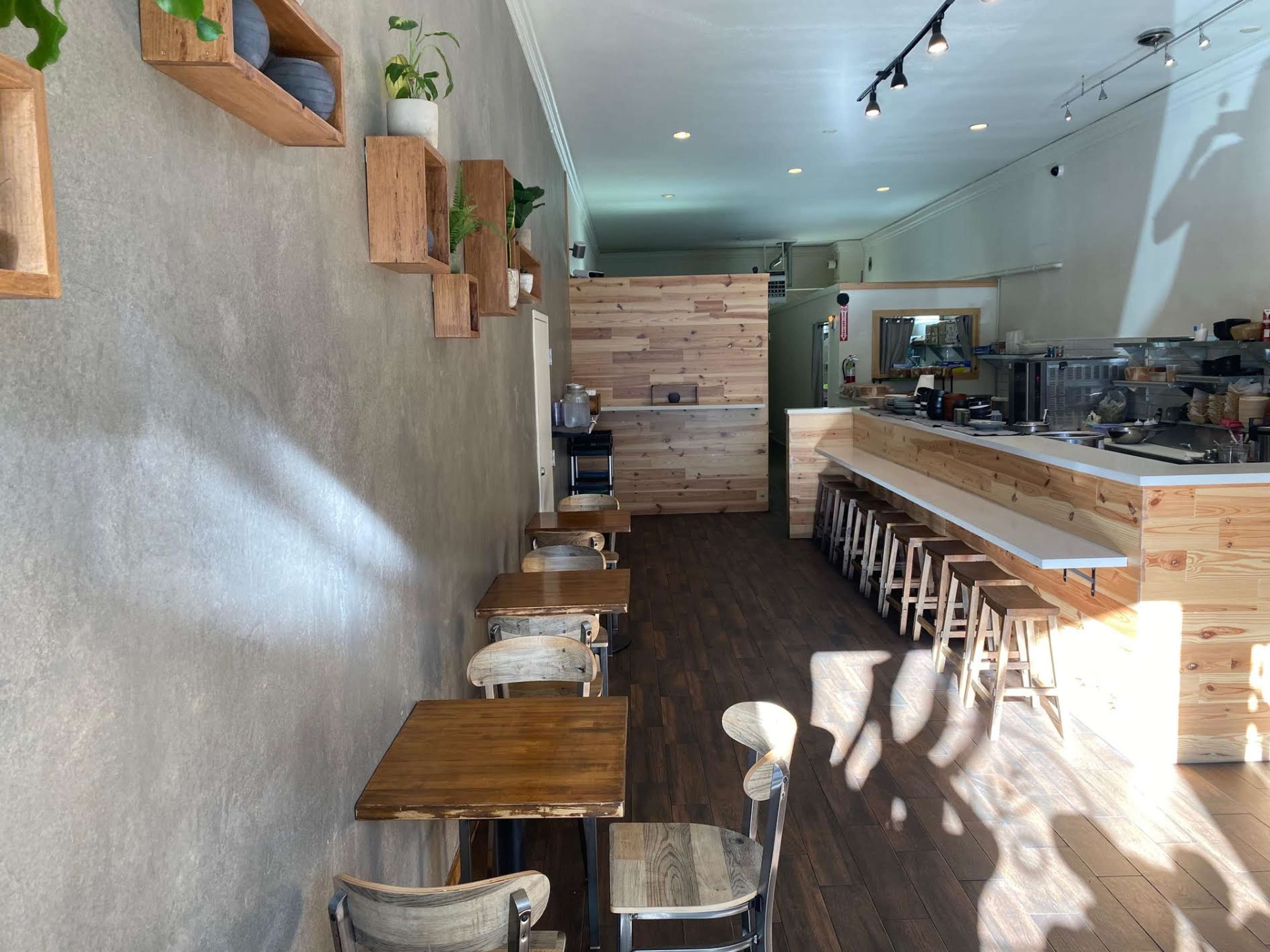 A minimalist café interior with wooden tables, high stools, and a light-colored wall adorned with planters.