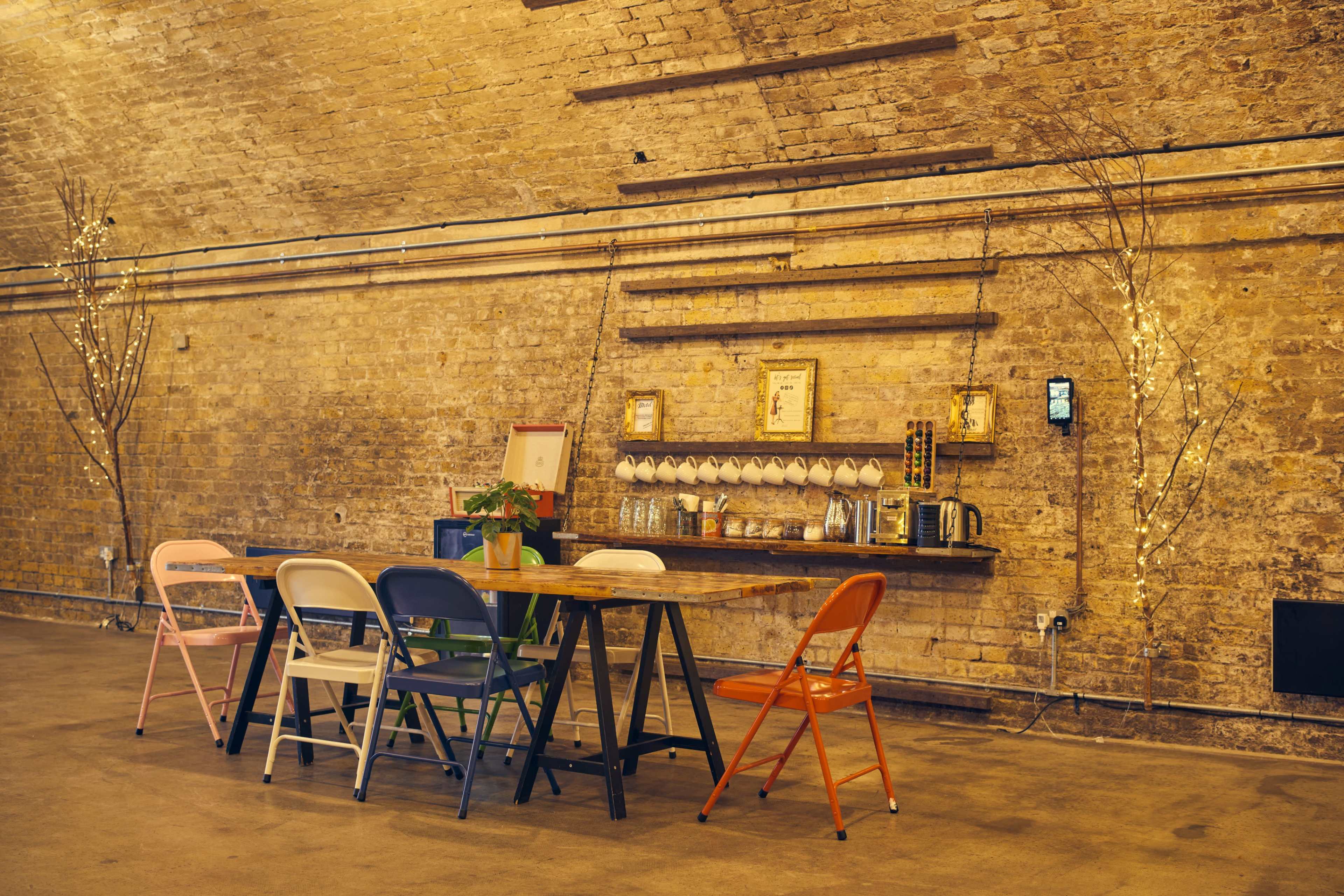 A long wooden table surrounded by colorful chairs is set against a textured brick wall with decorative lighting and a shelf displaying various items.