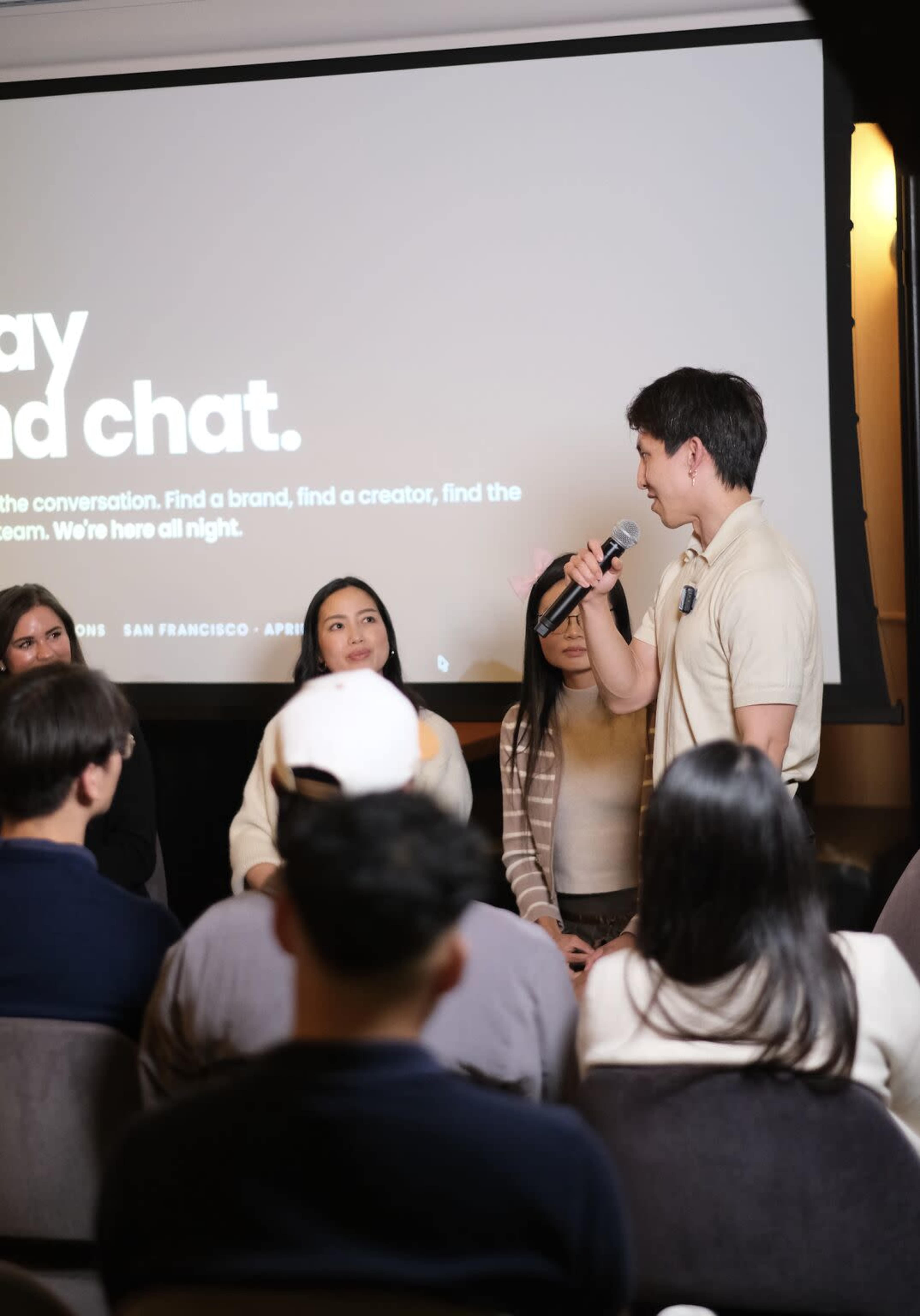 A speaker addresses an audience during an event, with a presentation screen in the background displaying the text "Let's play and chat."
