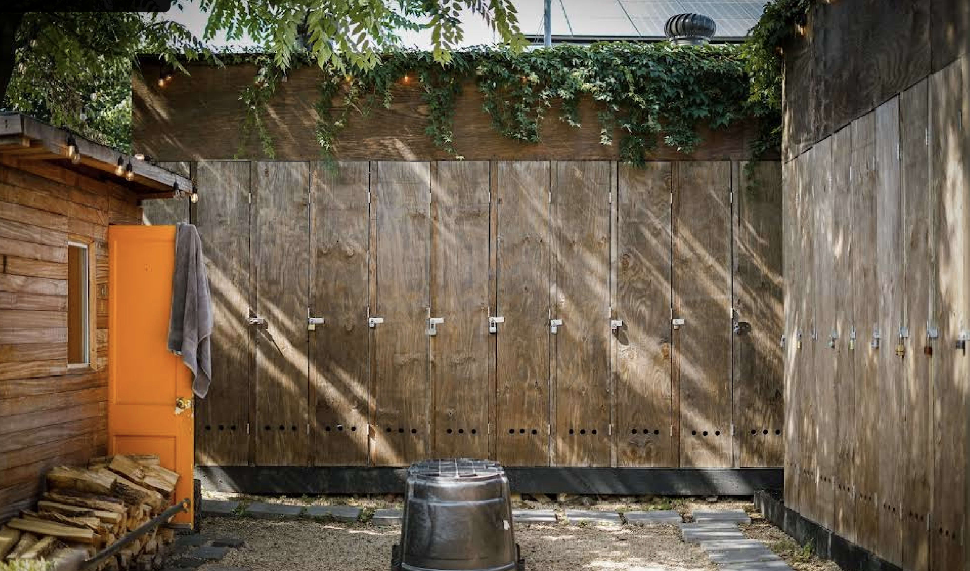 The image shows an outdoor area with wooden lockers against a wall, surrounded by greenery and a round metal container on the ground.