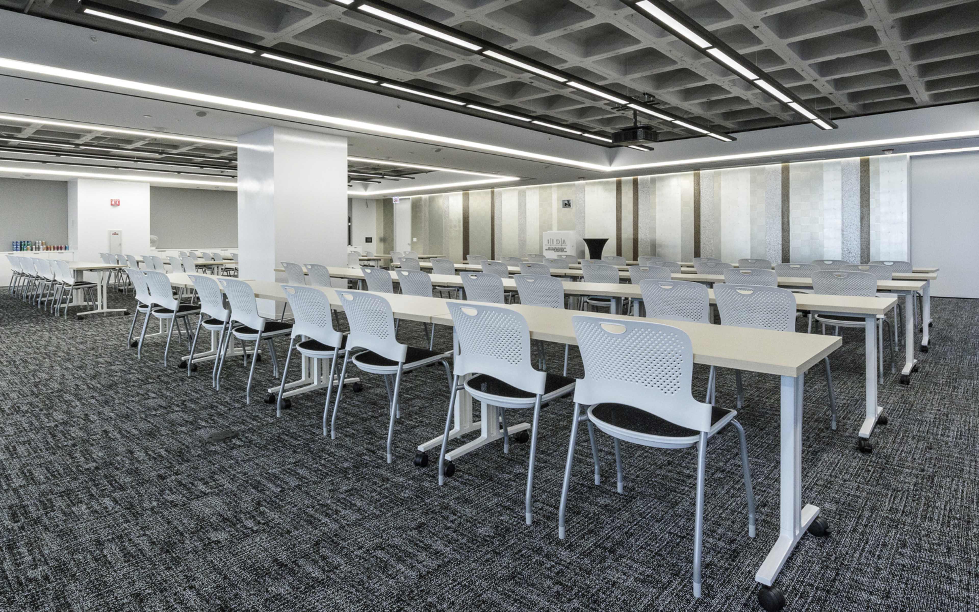 A large, modern conference room features multiple rows of tables and chairs arranged neatly on a carpeted floor.