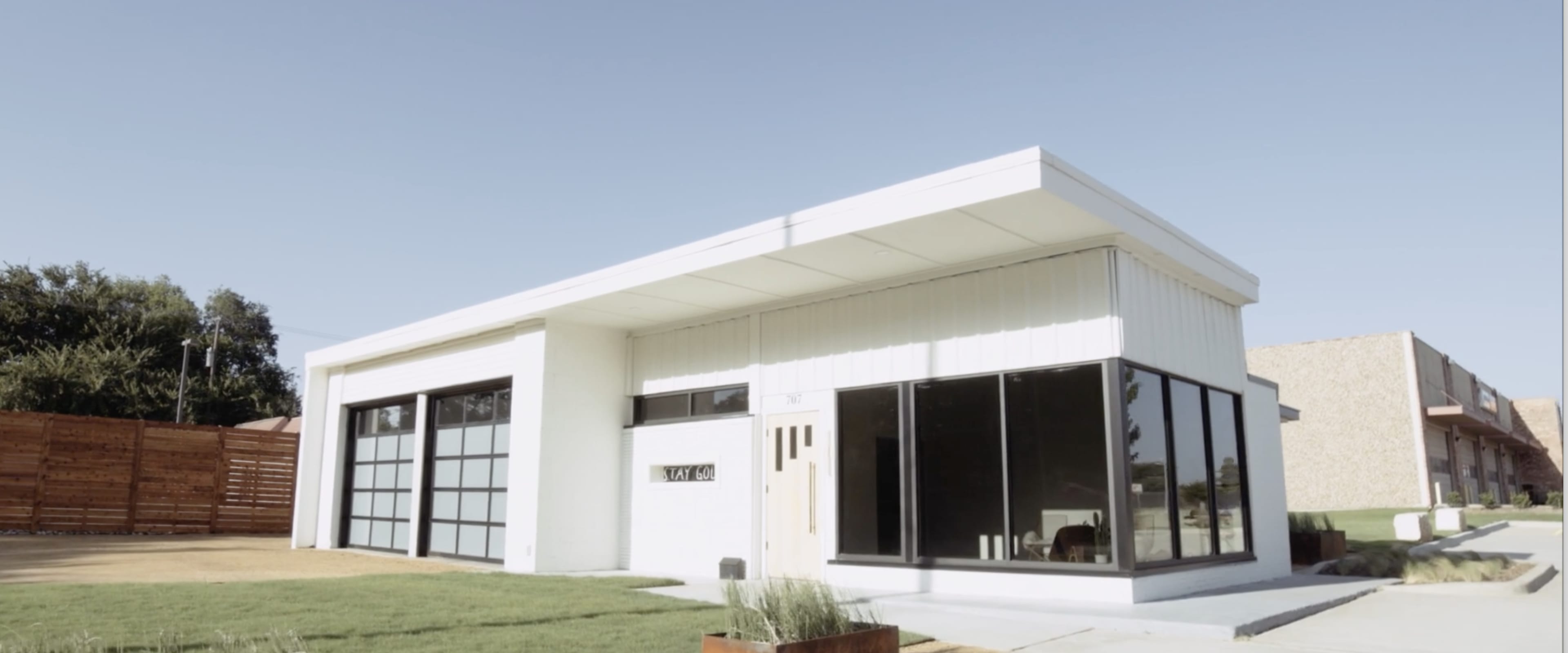 A modern single-story house with large glass windows and a flat roof, surrounded by a lawn and a gravel path.