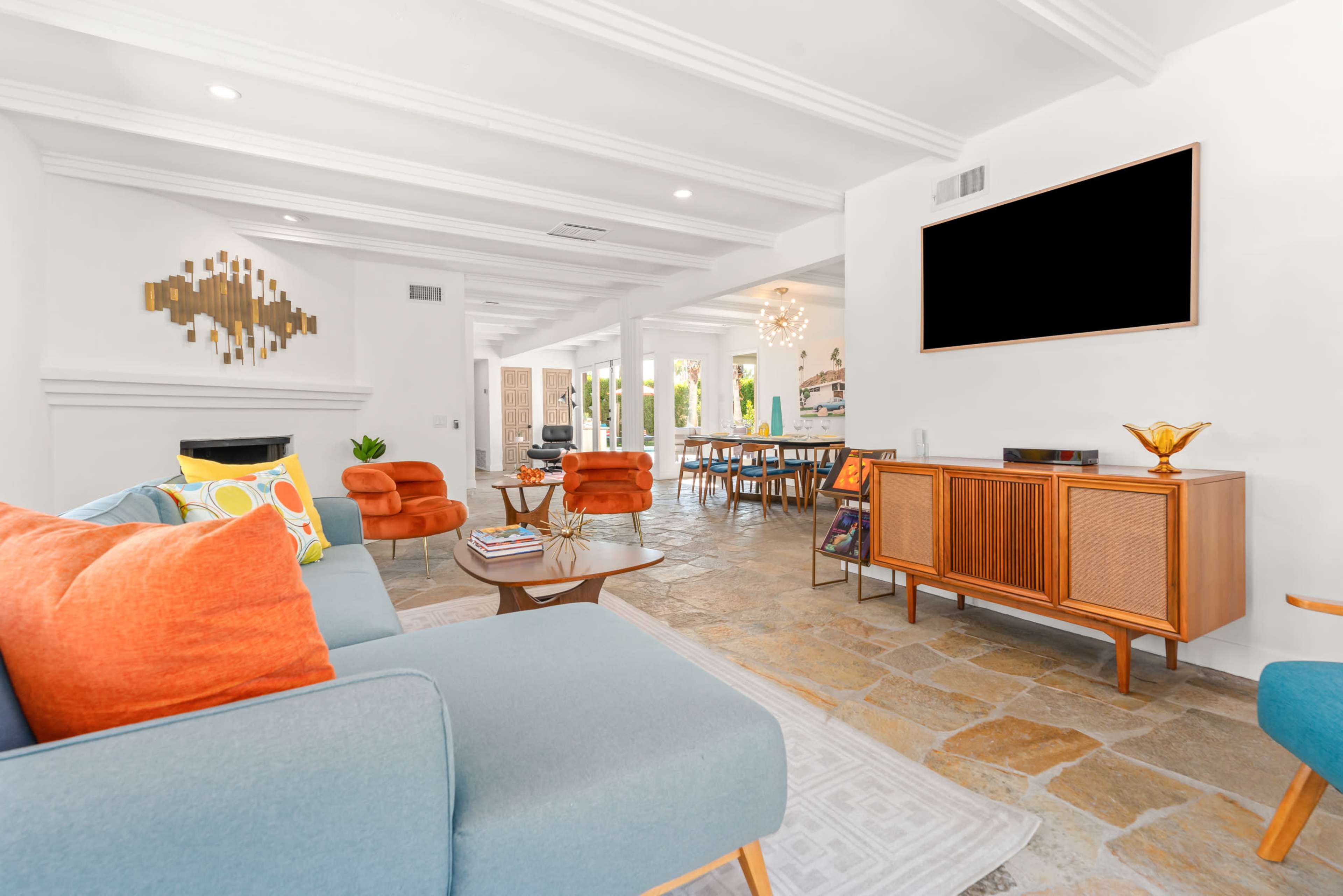 The image shows a modern living room with a blue couch, orange armchairs, a wooden media console, and a dining area visible in the background.