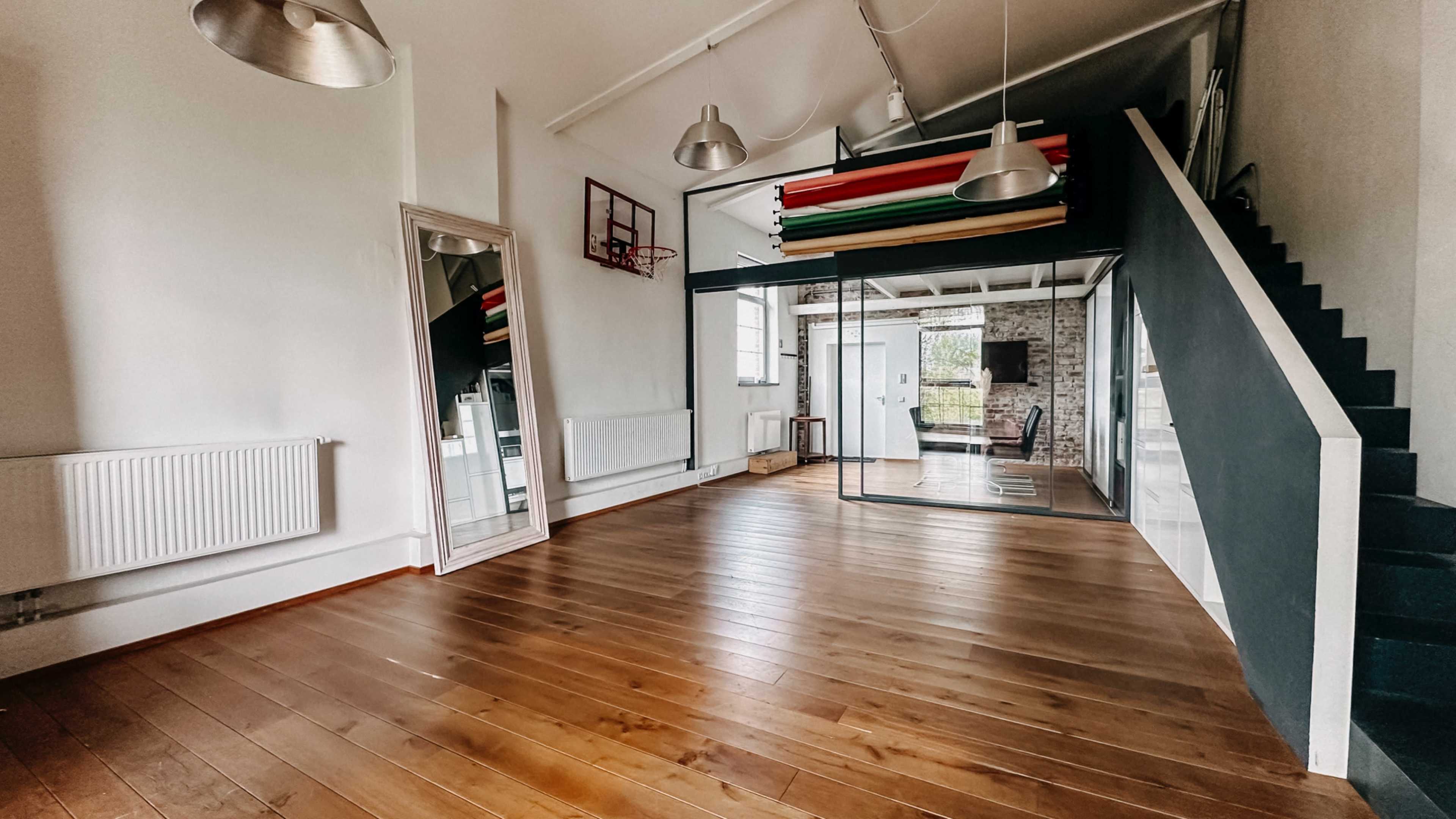 The image shows a spacious room with wooden flooring, large windows, and colorful gym mats displayed on a shelf.